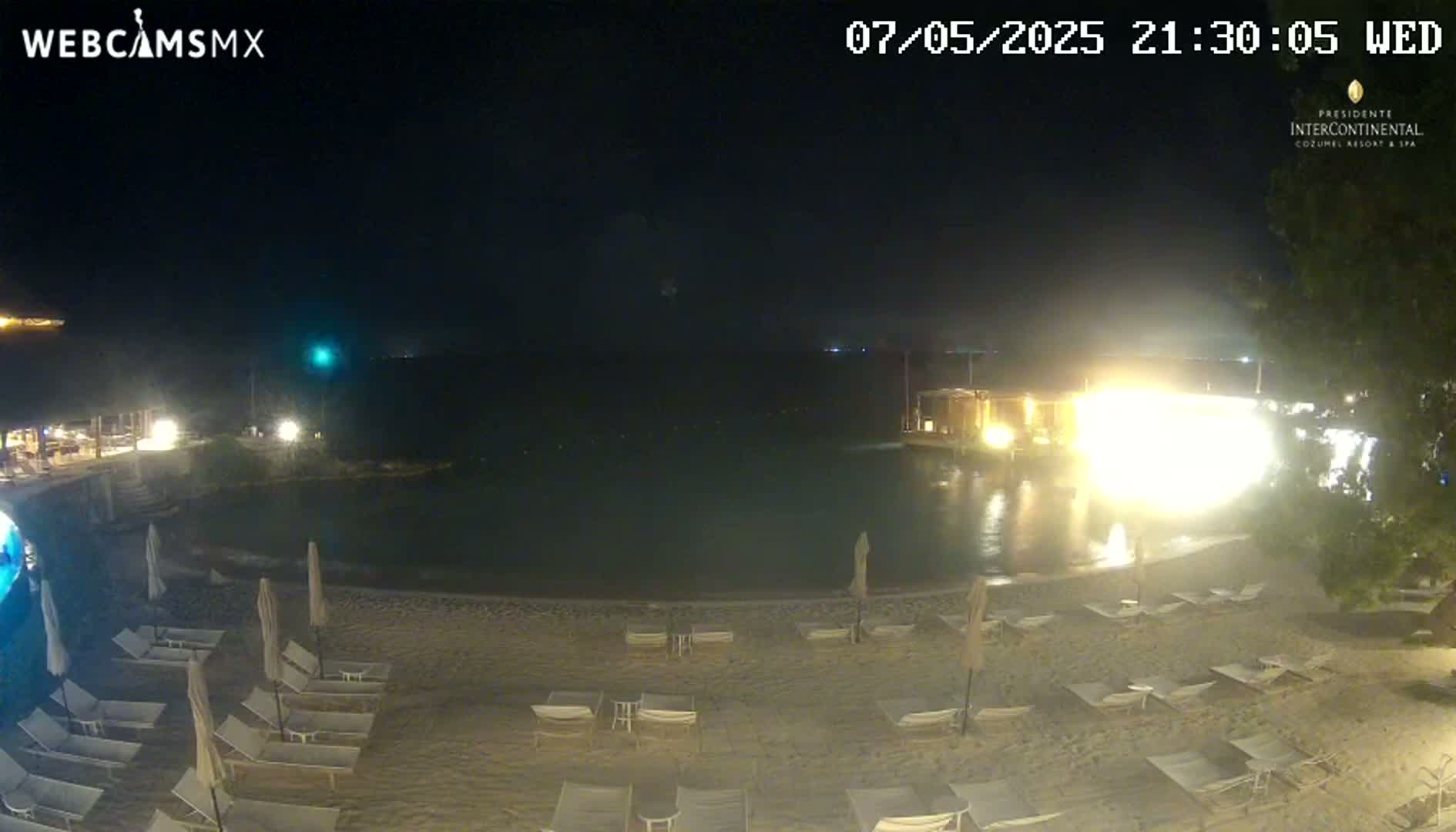 A nighttime beach scene shows a brightly lit building near a calm body of water, with lounge chairs and umbrellas set up on a sandy shore.