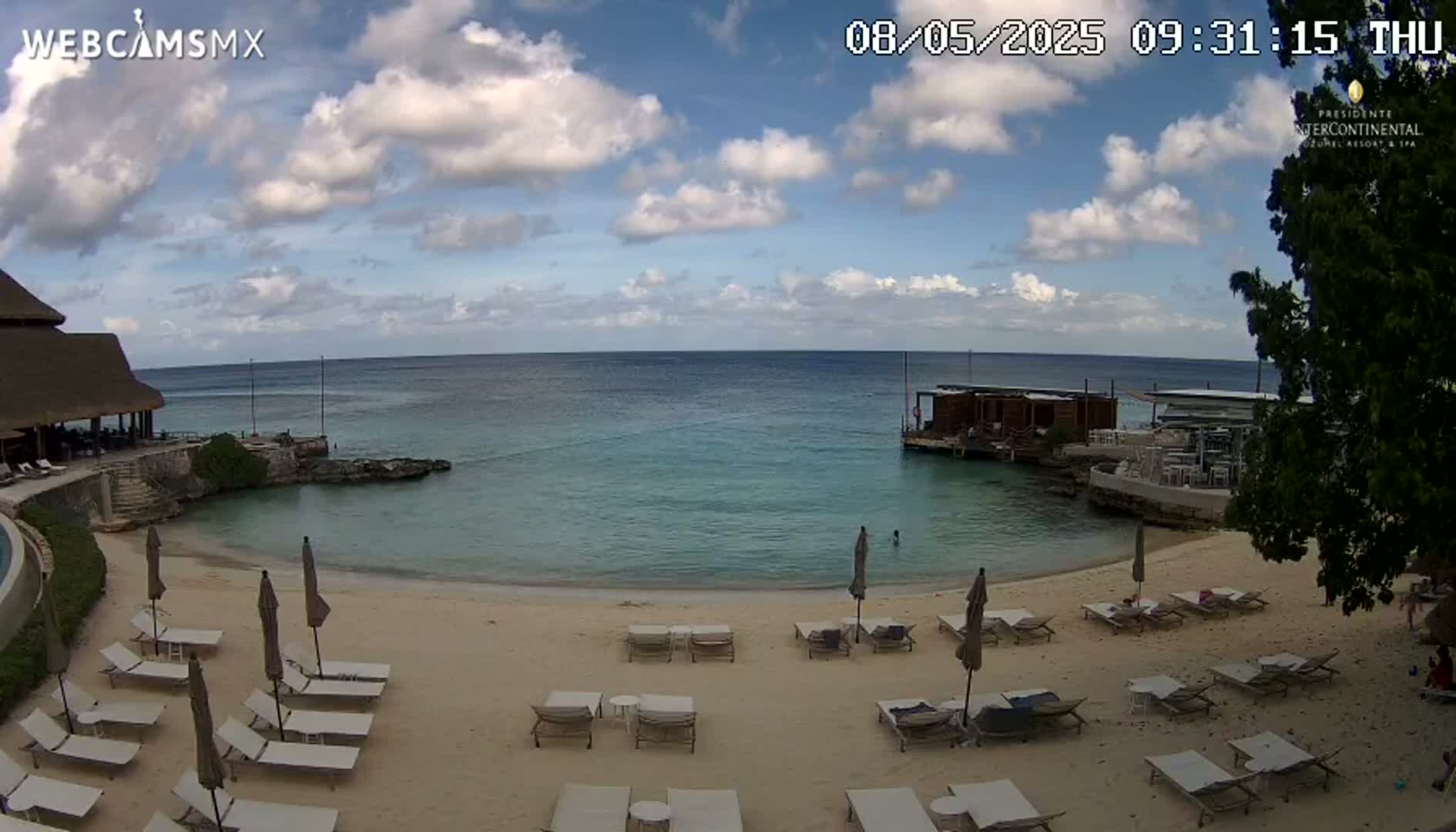 A partly cloudy day shows a sandy beach with lounge chairs and umbrellas, a calm ocean, and a small overwater structure.
