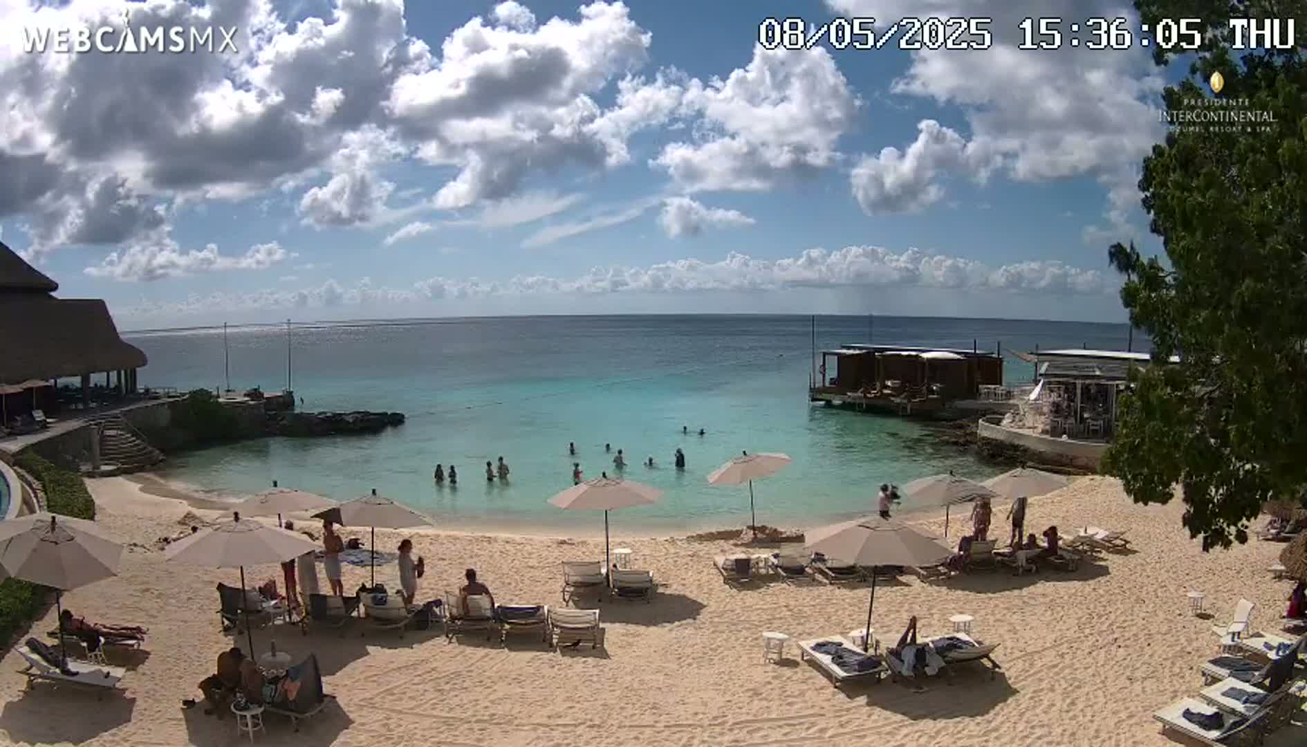 A partly cloudy sunny day at a beach with calm, shallow turquoise water, several people swimming and sunbathing on a sandy beach with lounge chairs and umbrellas.