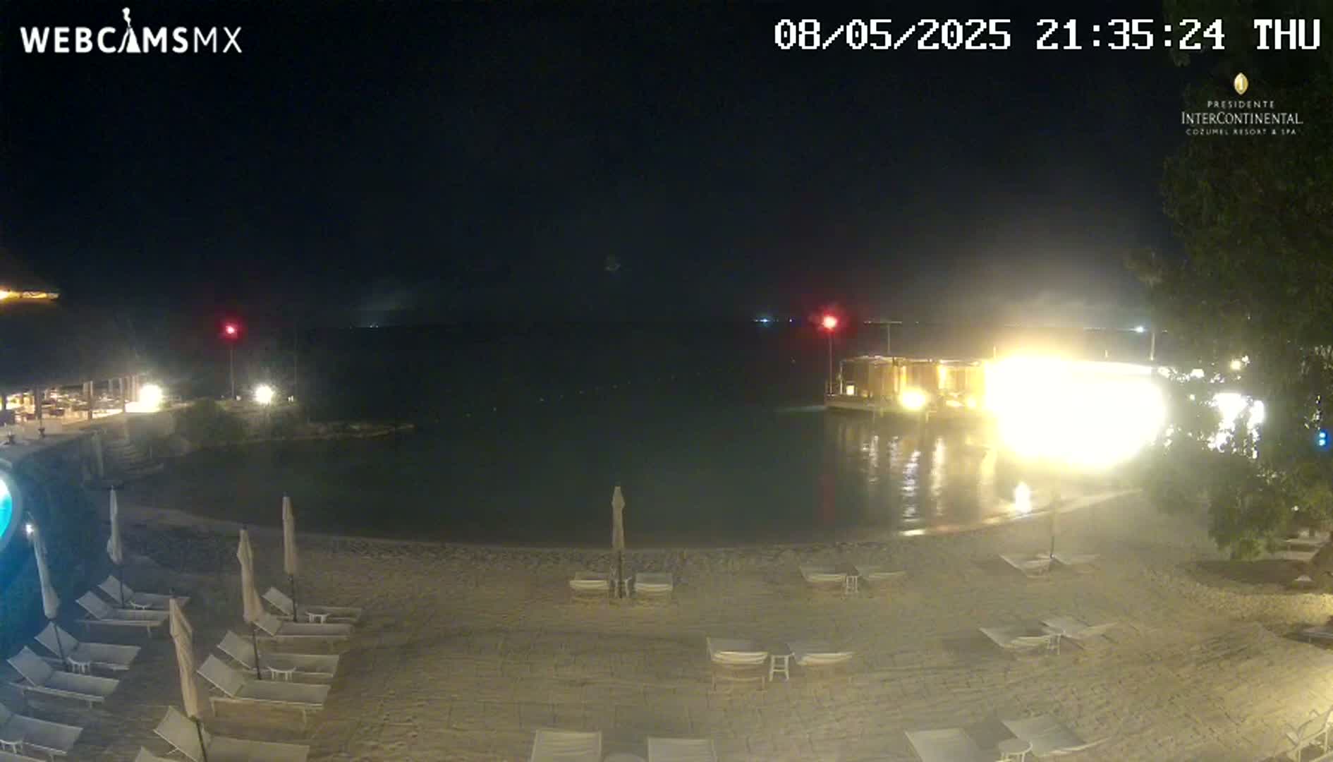 A nighttime view of a beach with lounge chairs and umbrellas, a calm body of water, and brightly lit buildings in the background.