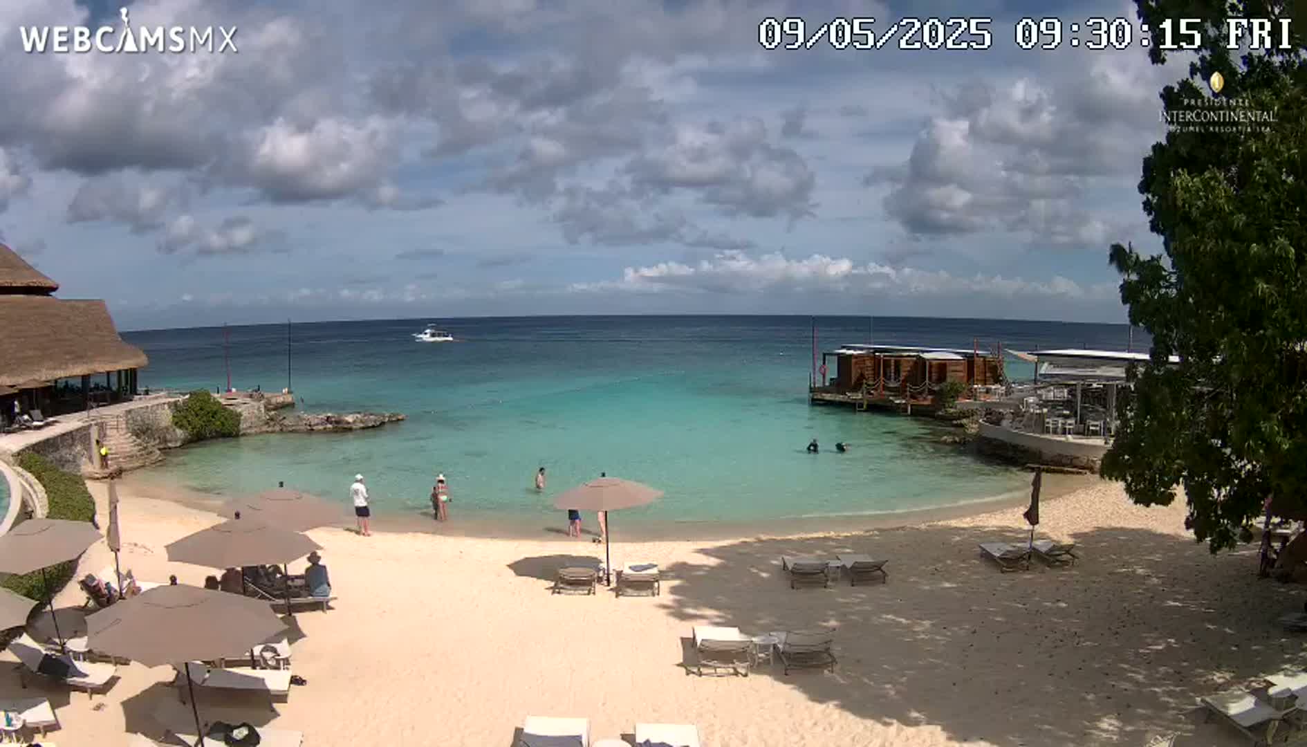 A partly cloudy day shows a beach with lounge chairs and umbrellas, calm turquoise water, and a few people swimming and relaxing.