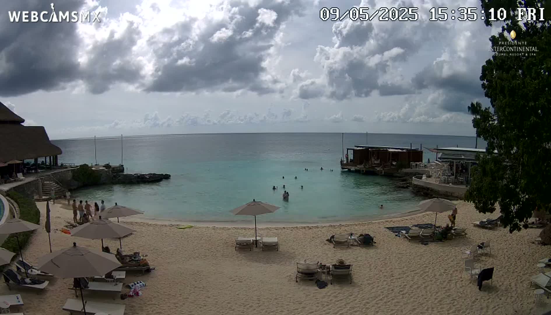 A partly cloudy day at a beach with turquoise water, people swimming and sunbathing on the sand under beach umbrellas and lounge chairs near a resort.