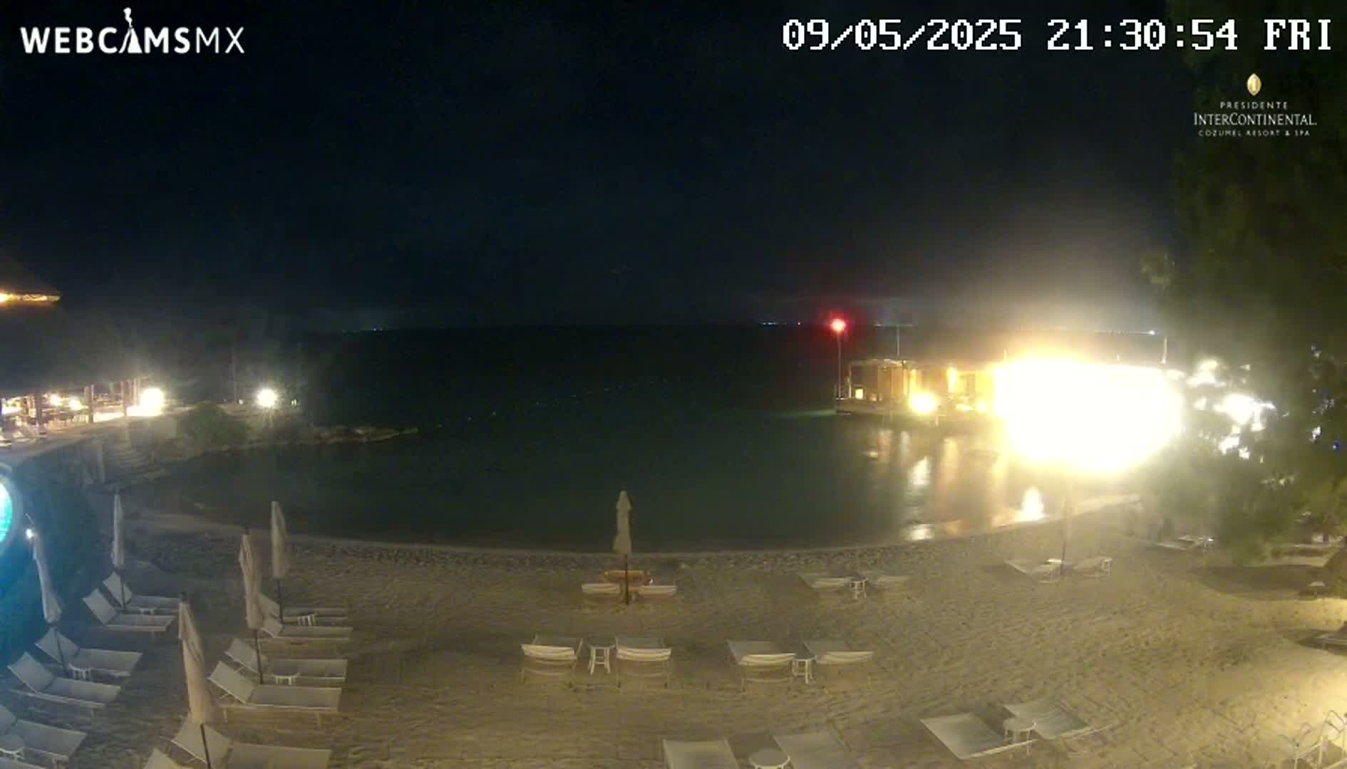 A nighttime beach scene shows calm water, a sandy shore with lounge chairs and umbrellas, and brightly lit buildings in the background.