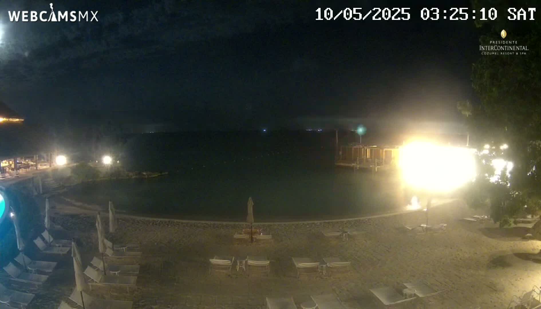 A nighttime beach scene shows a calm body of water, a sandy shoreline with lounge chairs, and bright lights illuminating a nearby structure.