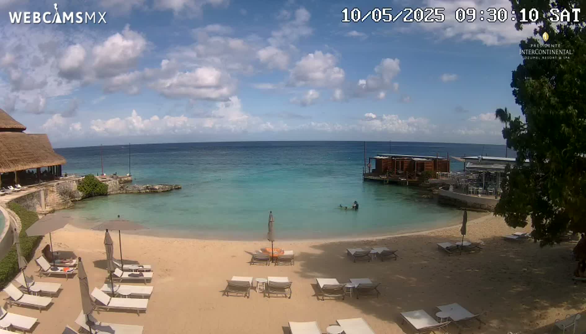 A sandy beach with lounge chairs and umbrellas sits next to calm, turquoise water under a partly cloudy sky.