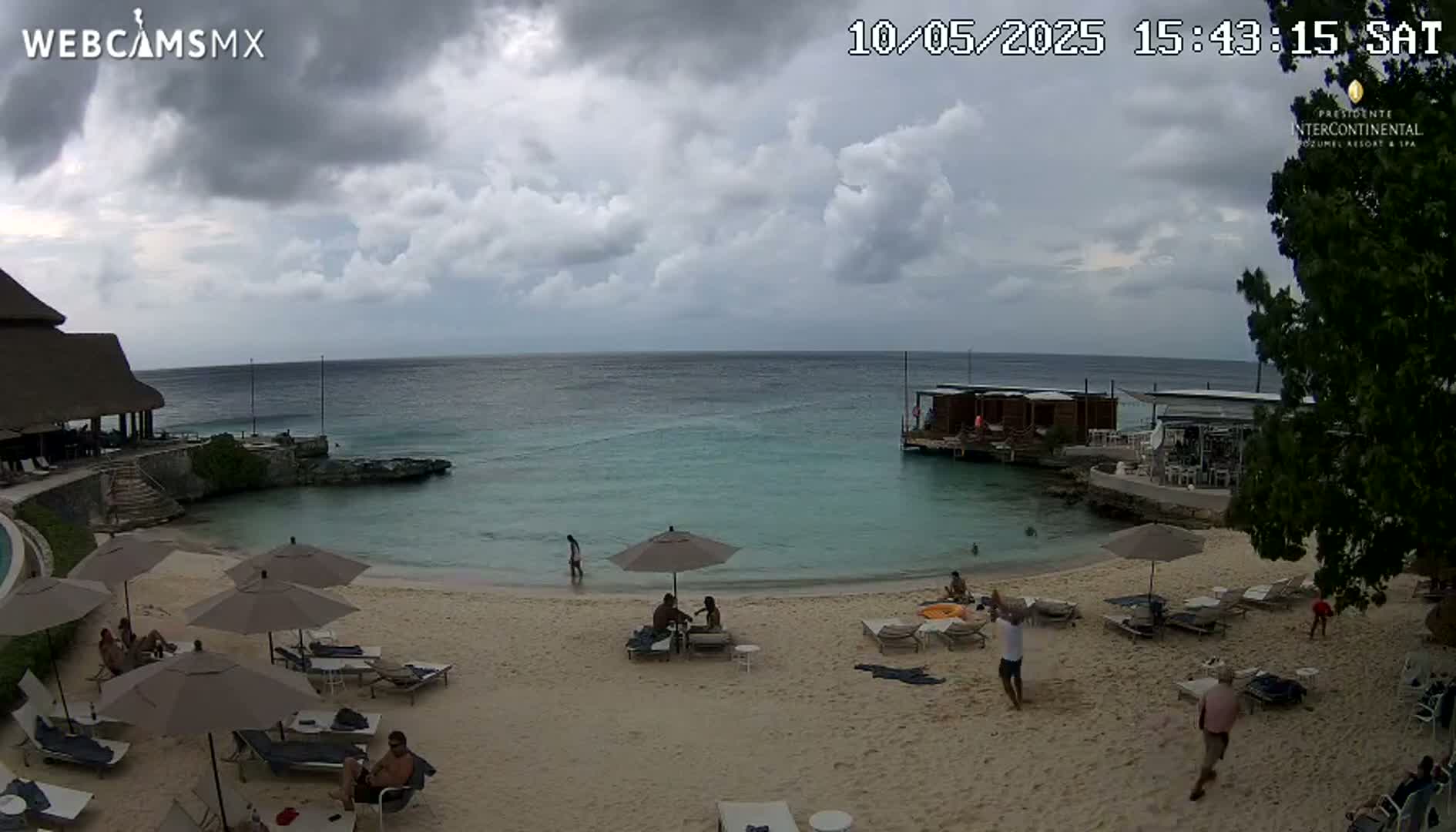 A beach scene under a partly cloudy sky shows people relaxing under umbrellas on a sandy beach near calm, turquoise water and a shoreline with buildings.