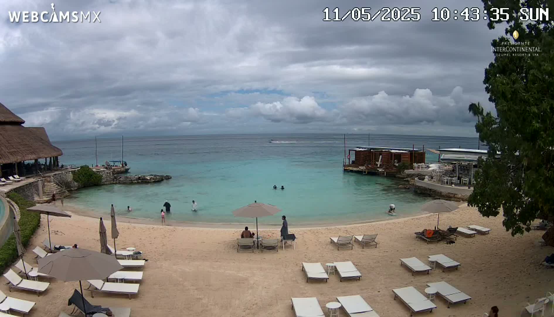 A partly cloudy day at a beach with calm, turquoise water, a sandy shore lined with lounge chairs and umbrellas, and a few people swimming and lounging.