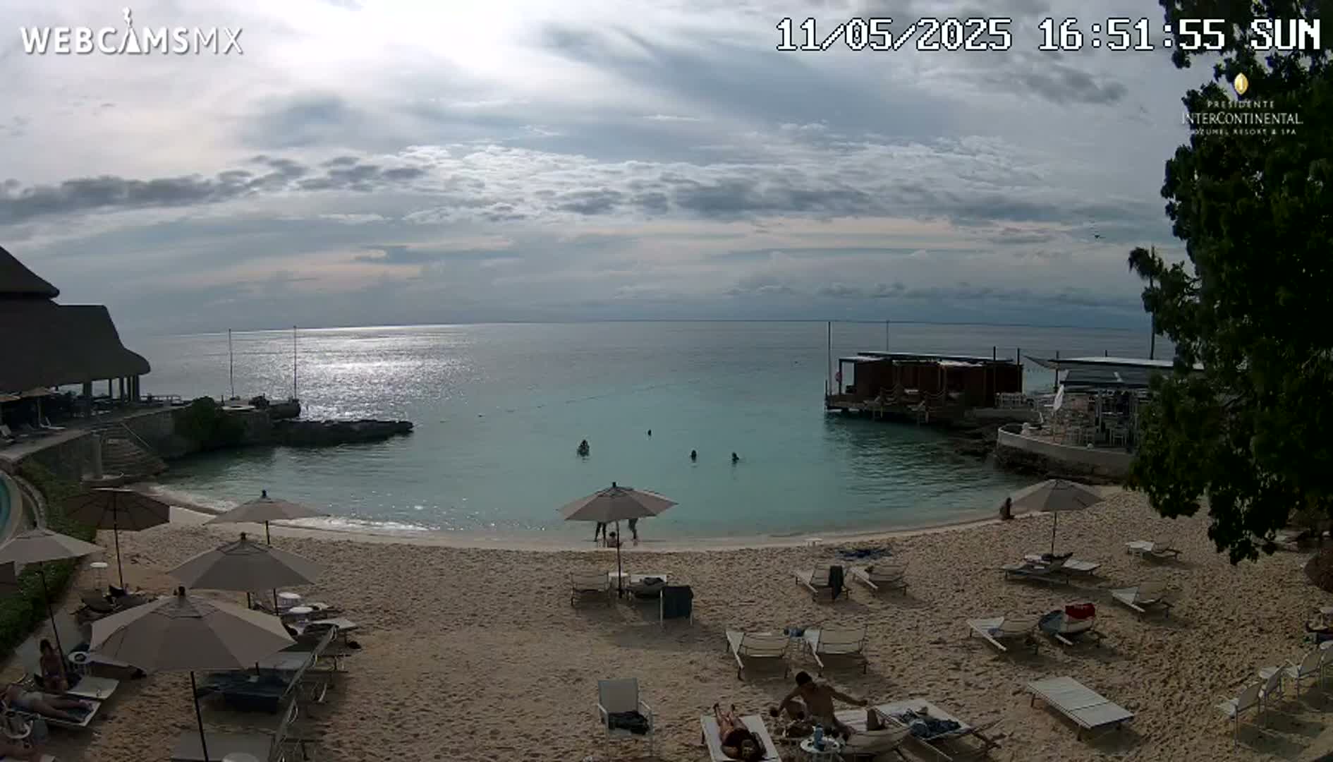 A partly cloudy day shows a sandy beach with lounge chairs and umbrellas, calm turquoise water, and a resort building with a thatched roof.
