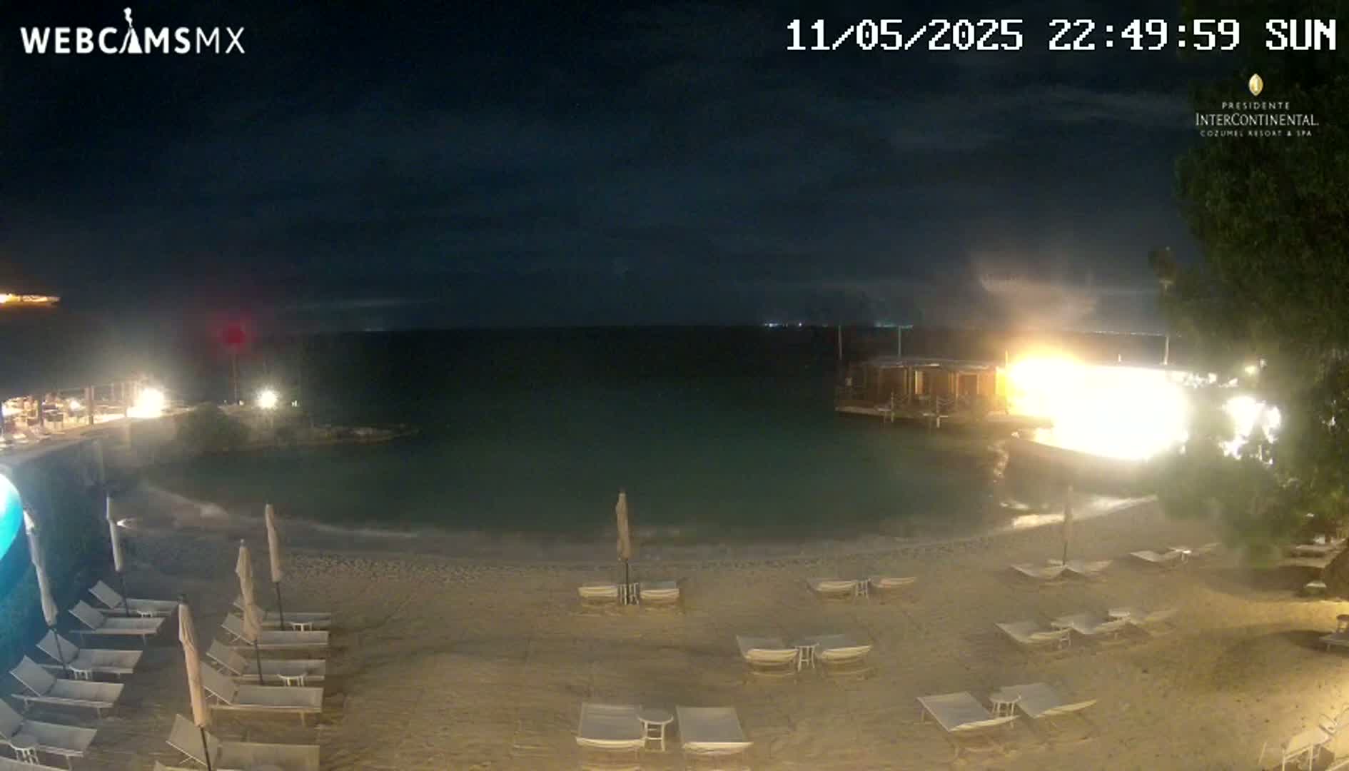 A nighttime beach scene shows a calm body of water, a sandy shore with lounge chairs, and bright lights illuminating a structure near the water.