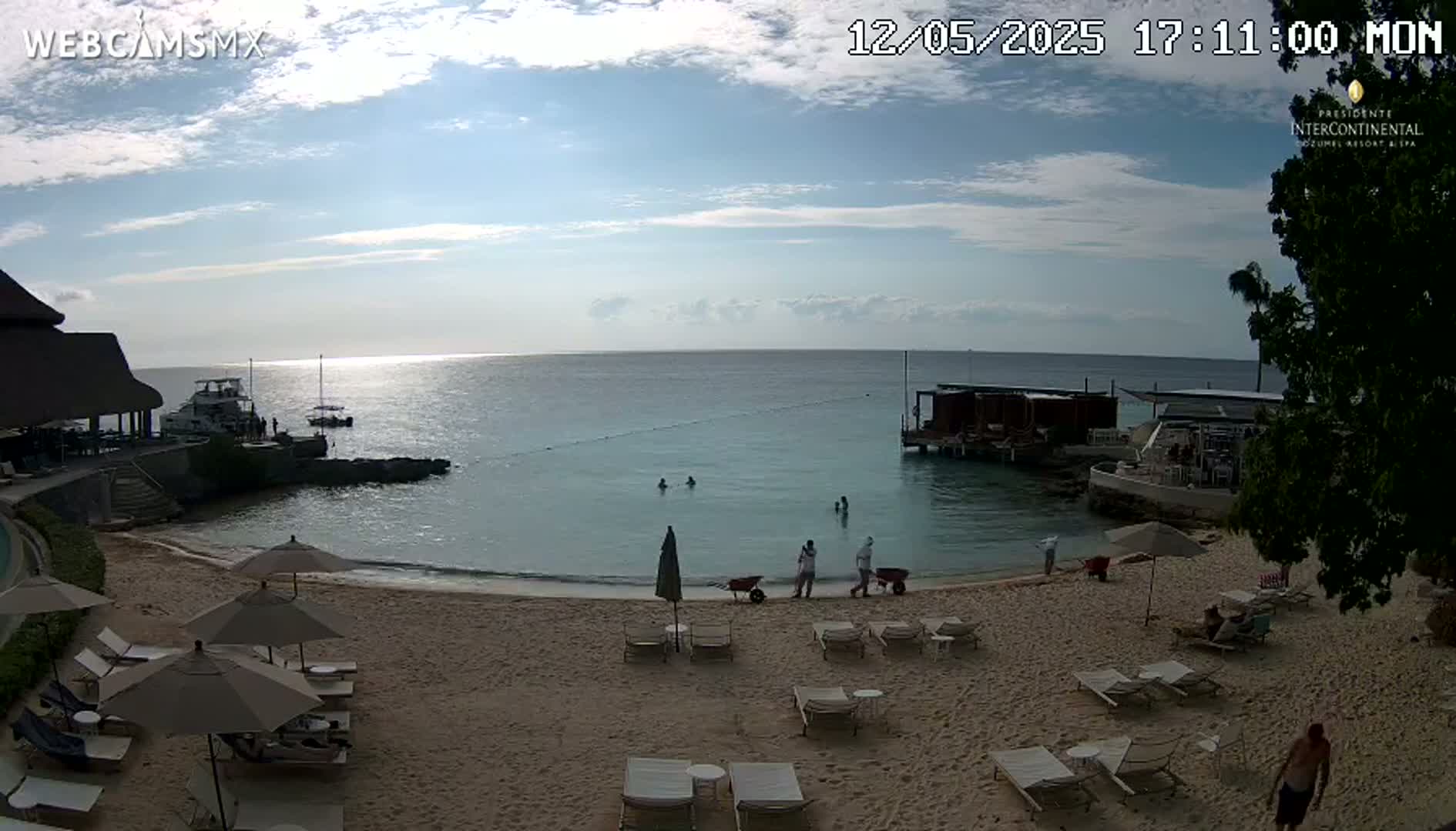 A sandy beach with lounge chairs and umbrellas is shown, with calm, clear ocean water in the background under a partly cloudy sky.