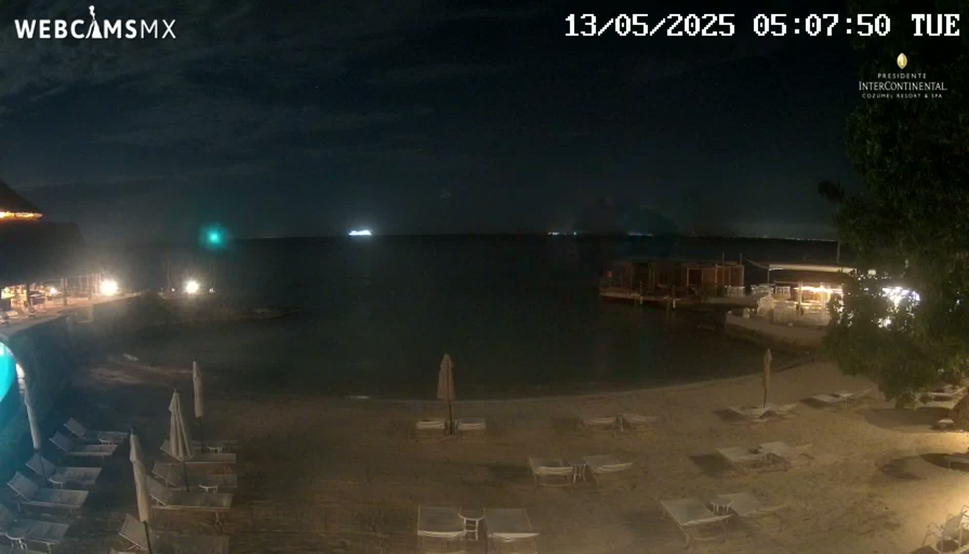 A nighttime view of a sandy beach with lounge chairs and umbrellas, overlooking a calm body of water with lights visible in the distance.