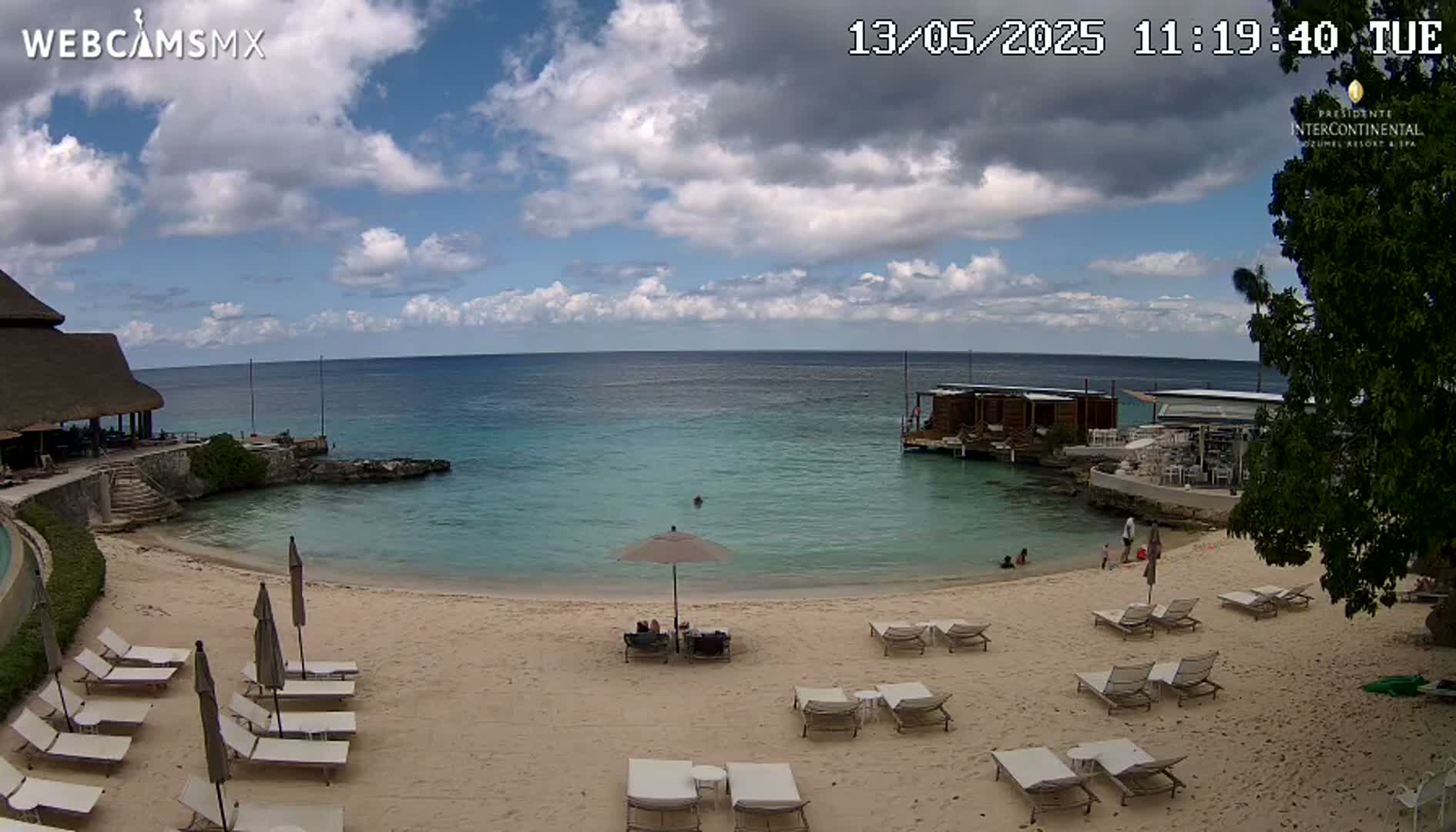 A partly cloudy day at a beach with calm, clear water, lounge chairs, and umbrellas on the sand, near a beachfront building.