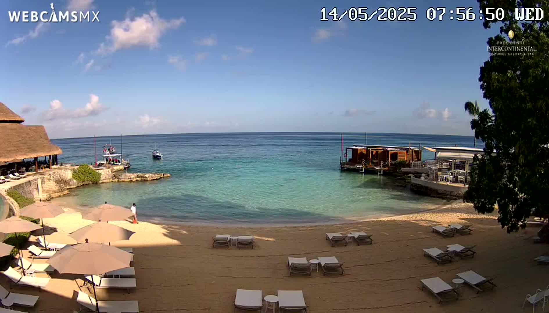 A sandy beach with lounge chairs and umbrellas sits on the shore of a calm, turquoise ocean under a mostly sunny sky.