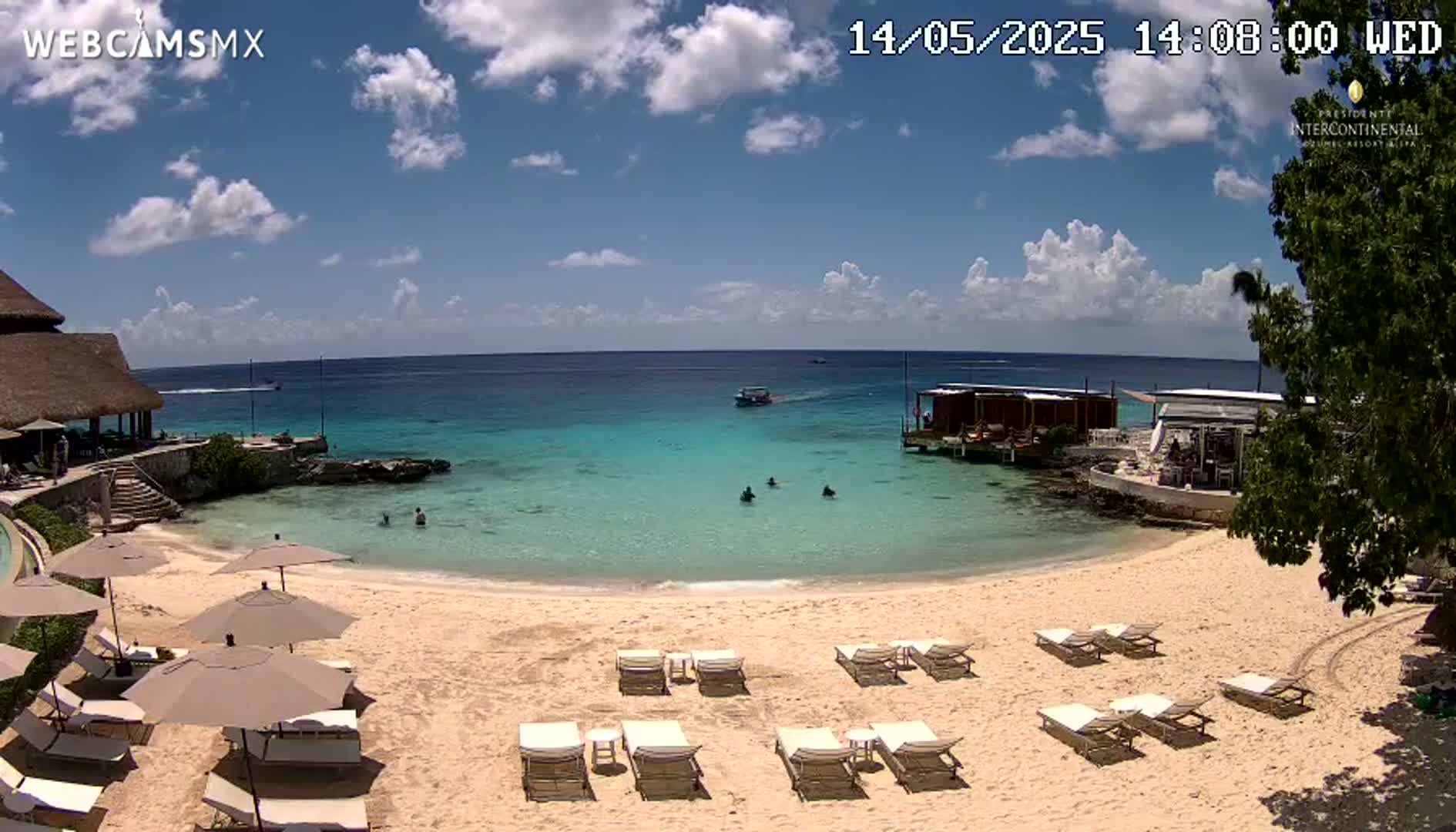 A partly sunny day shows a sandy beach with lounge chairs and umbrellas, calm turquoise water, and a resort building with a thatched roof.