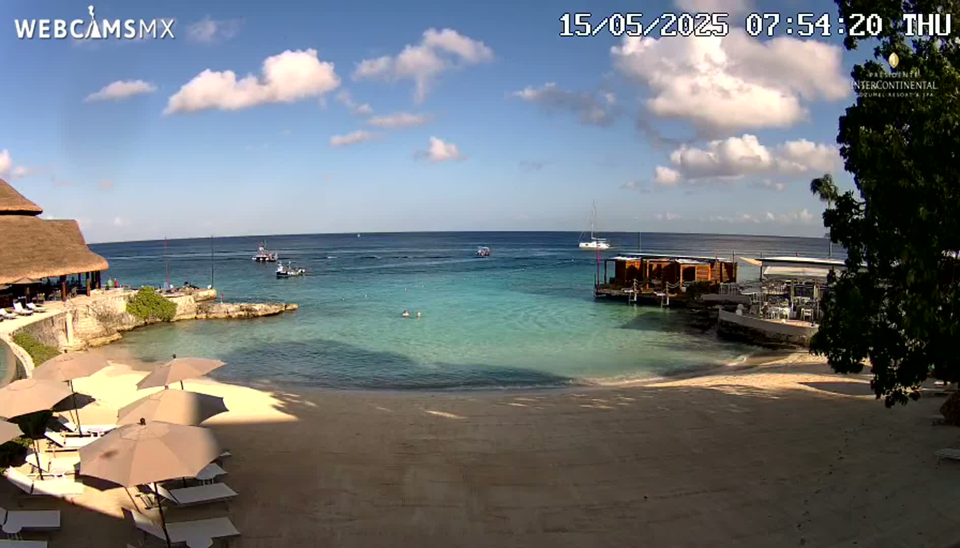 A partly sunny beach scene shows calm, clear turquoise water with several boats, beach umbrellas and lounge chairs, and a building with a thatched roof.