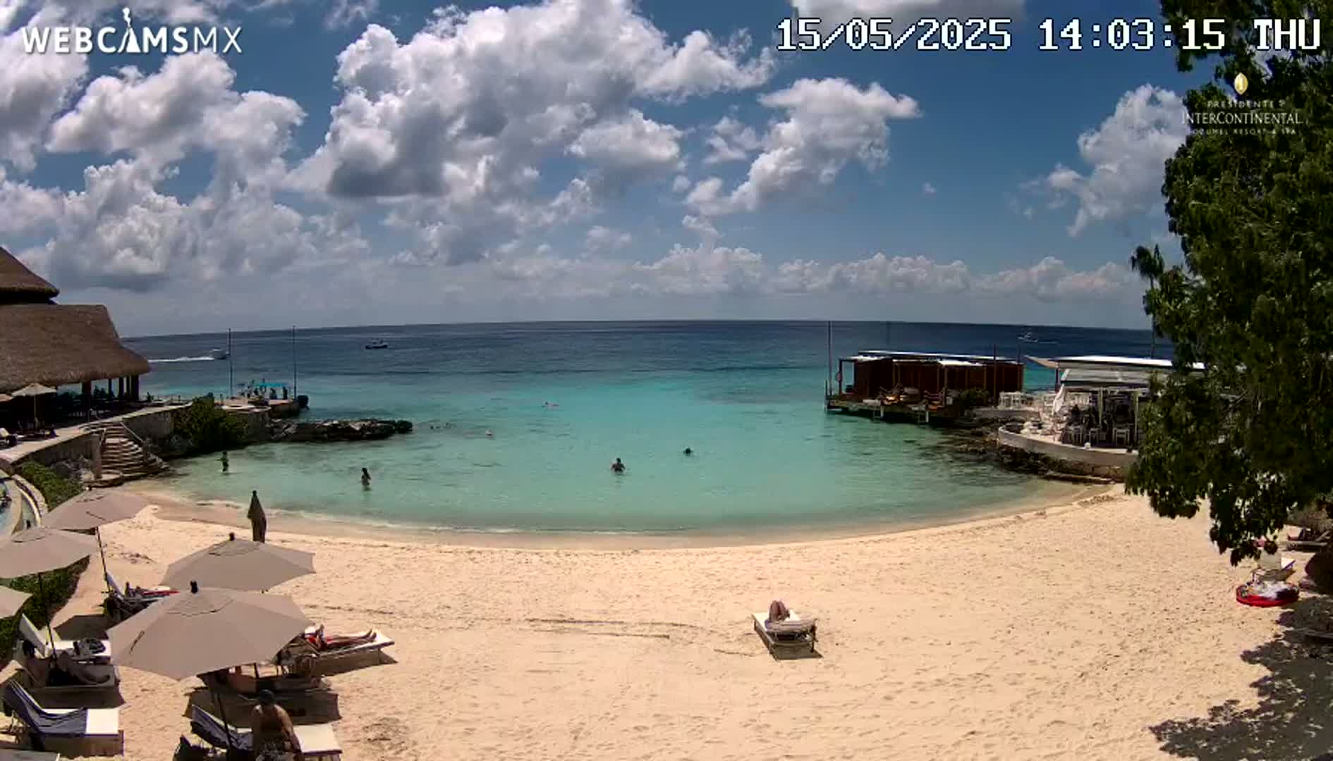 A partly cloudy day shows a sandy beach with calm, turquoise water, several people swimming, and beach umbrellas and loungers near a resort building.