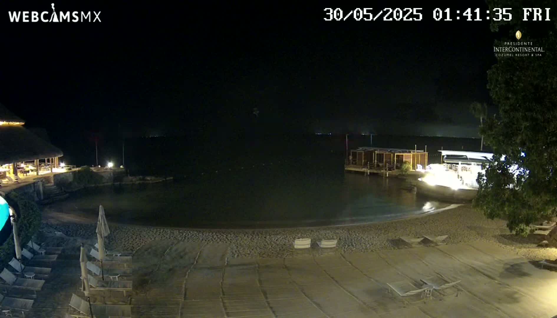 A nighttime view of a sandy beach with lounge chairs, a calm body of water, and buildings with lights visible in the distance.
