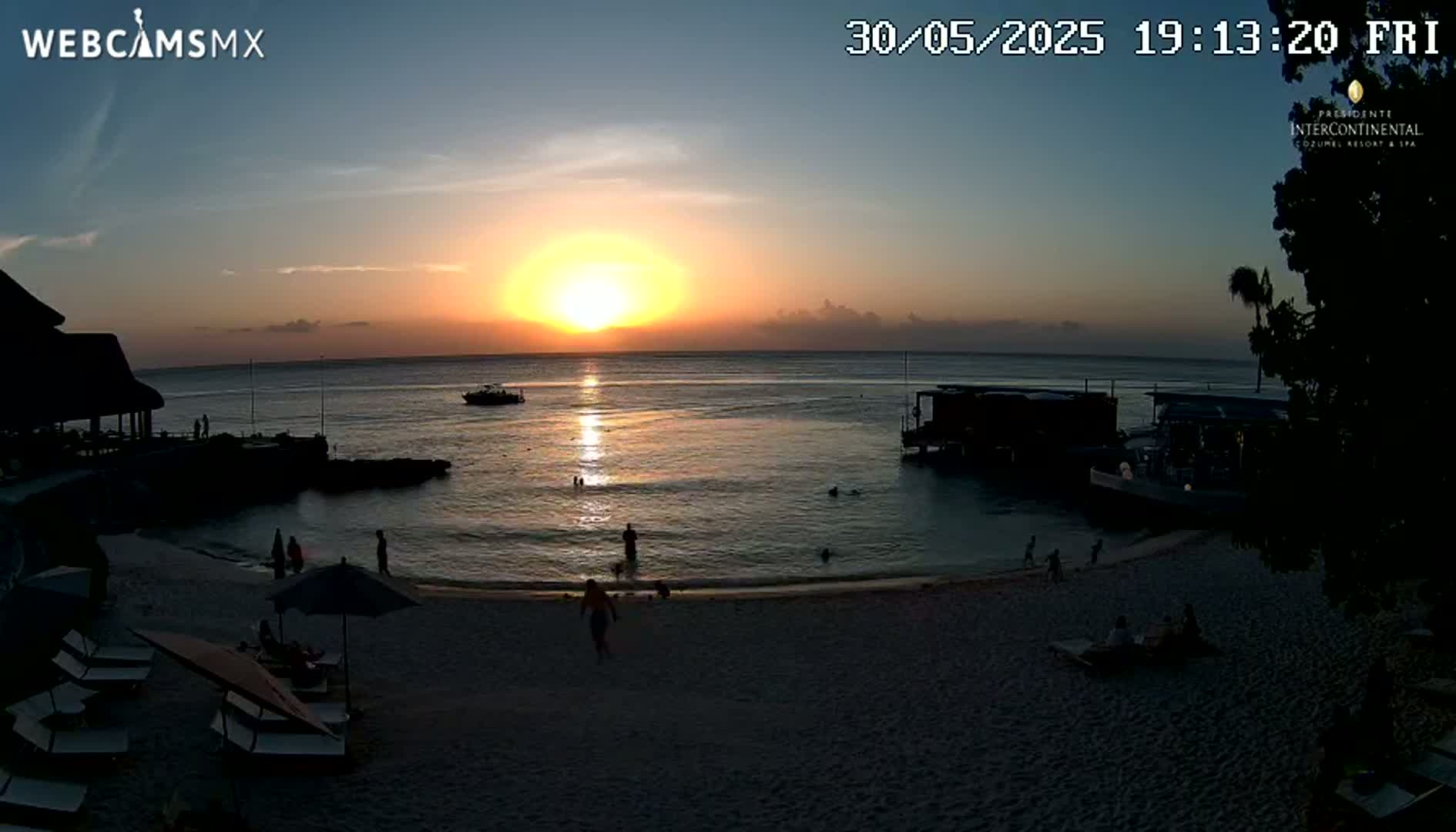 A beach scene at sunset shows a calm ocean with people swimming and lounging on the sand near beach umbrellas and lounge chairs, with buildings and boats visible in the background.