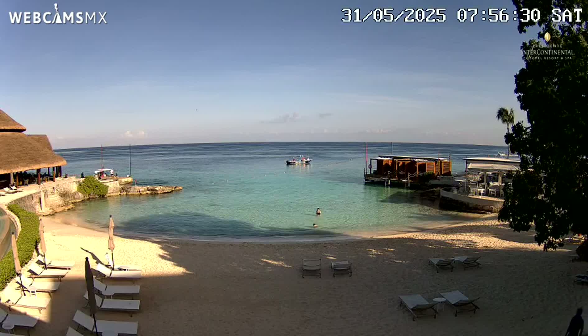 A sandy beach with lounge chairs and a calm, clear ocean under a sunny sky, featuring a small boat and an overwater structure.