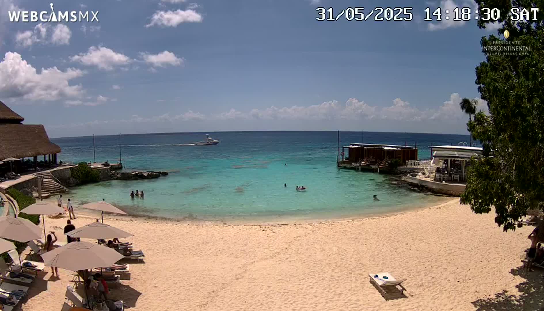 A sandy beach with turquoise water, several beach umbrellas, people swimming and relaxing, and a boat in the distance under a partly cloudy sky.