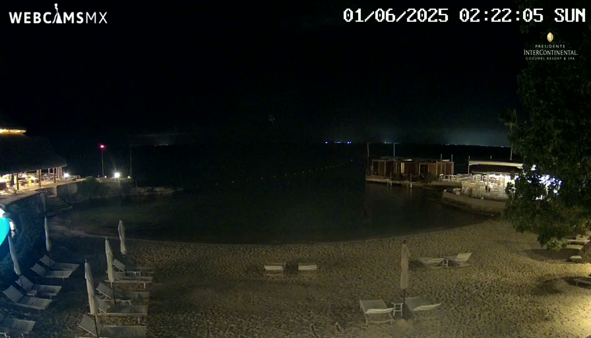 A nighttime beach scene shows a sandy shoreline with lounge chairs, umbrellas, and calm dark water under a clear, dark sky.