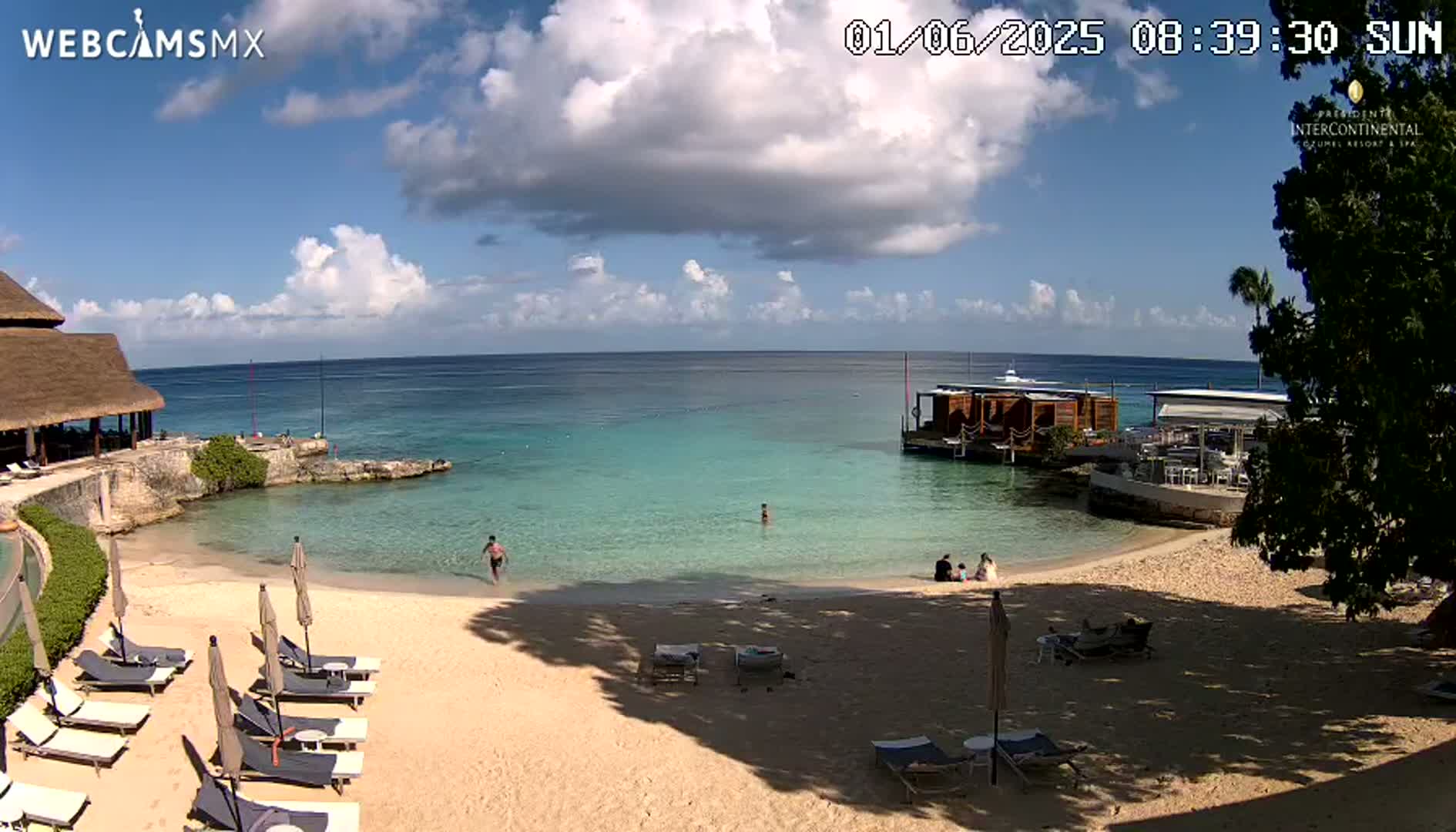 A partly sunny beach scene shows a calm turquoise ocean, a sandy beach with lounge chairs and umbrellas, a few people swimming and relaxing, and a building with a thatched roof.