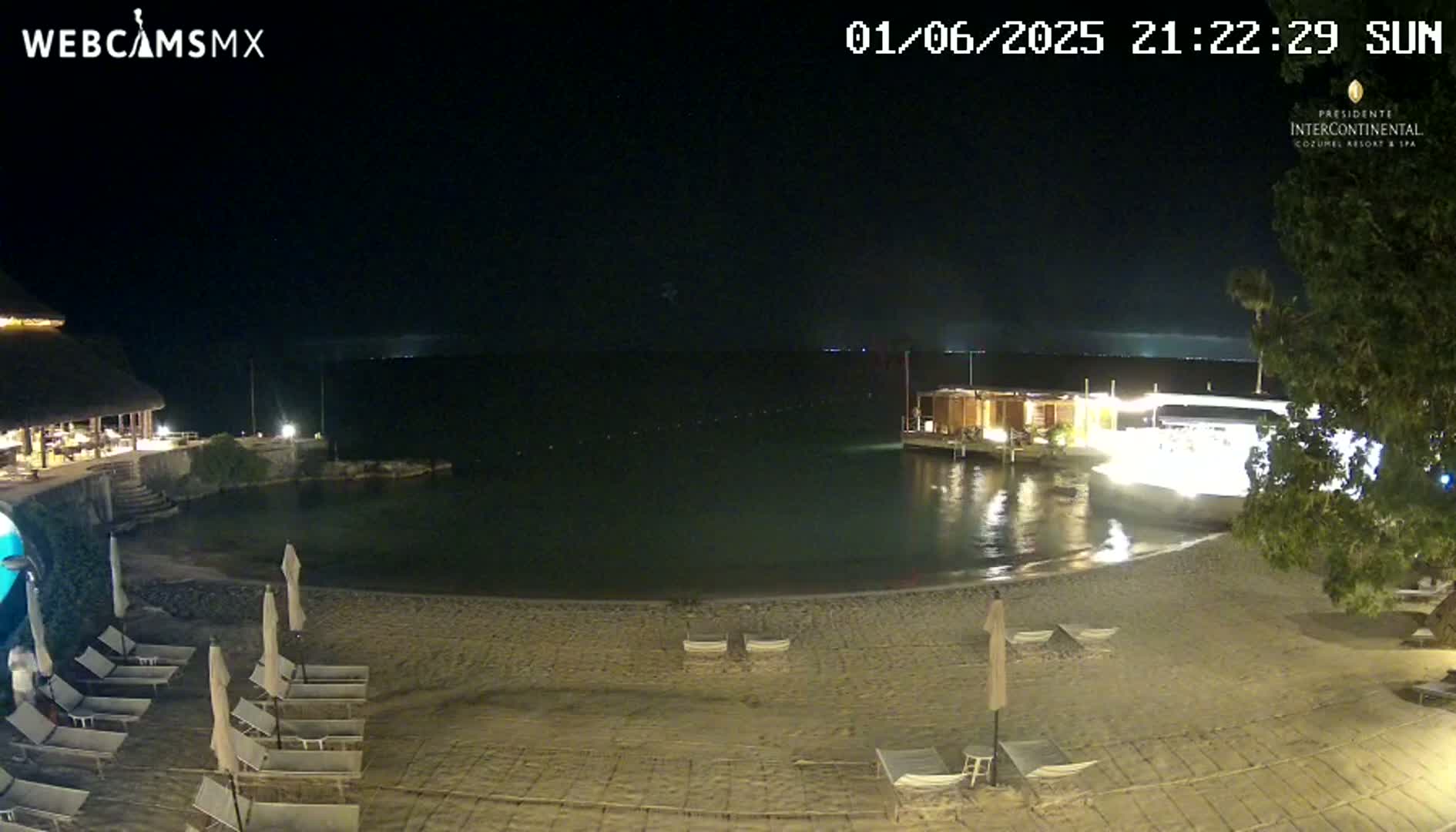 A nighttime beach scene shows a calm body of water, a sandy shore with lounge chairs and umbrellas, and a building with lights on in the background under a clear, dark sky.