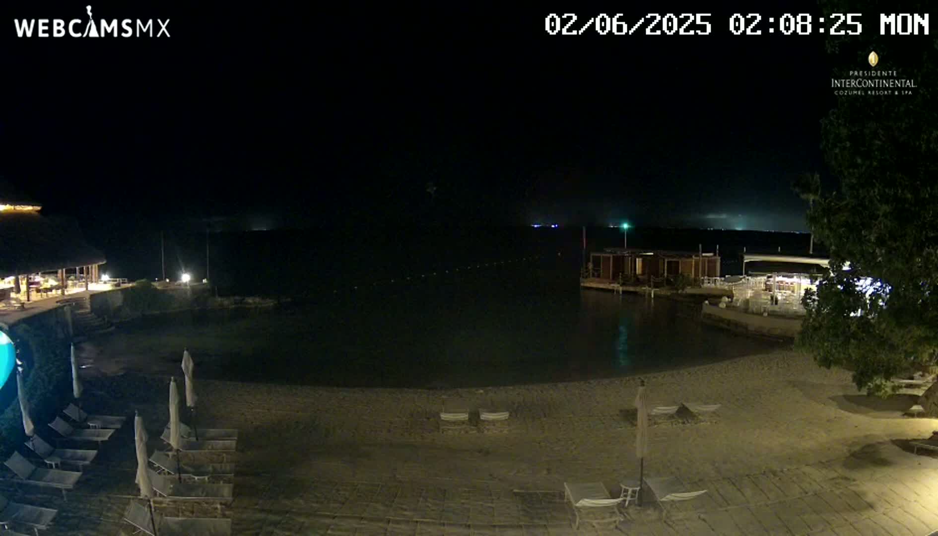 A nighttime beach scene shows a sandy shore with lounge chairs under an overcast sky, a calm body of water, and some resort buildings in the background.