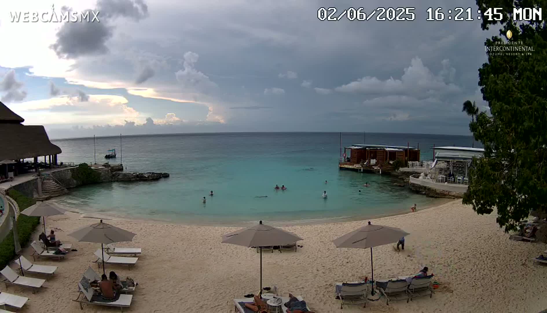 A partly cloudy day at a beach with calm, turquoise water, several people swimming and sunbathing on the sand, and beach umbrellas set up.