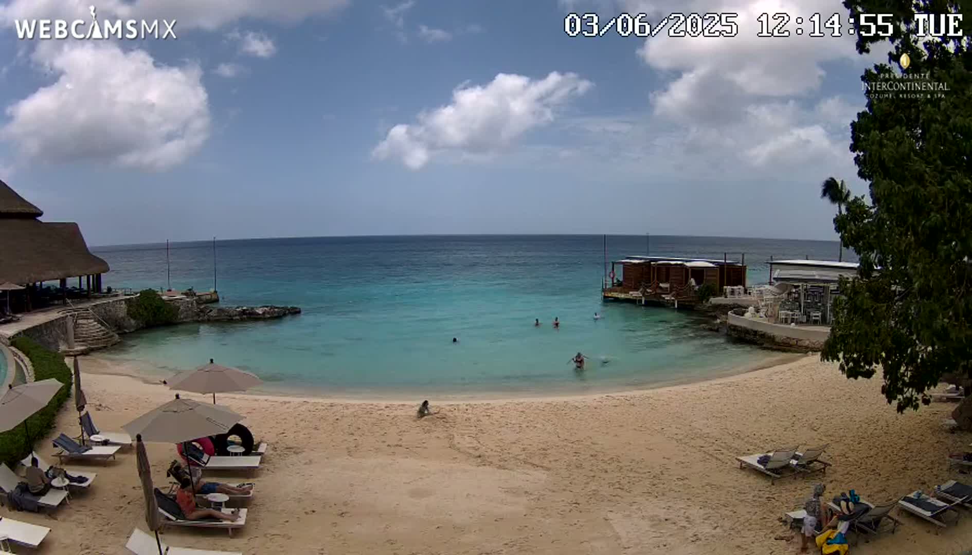 A sandy beach with turquoise water, a few people swimming, and lounge chairs under umbrellas is visible on a partly cloudy day.