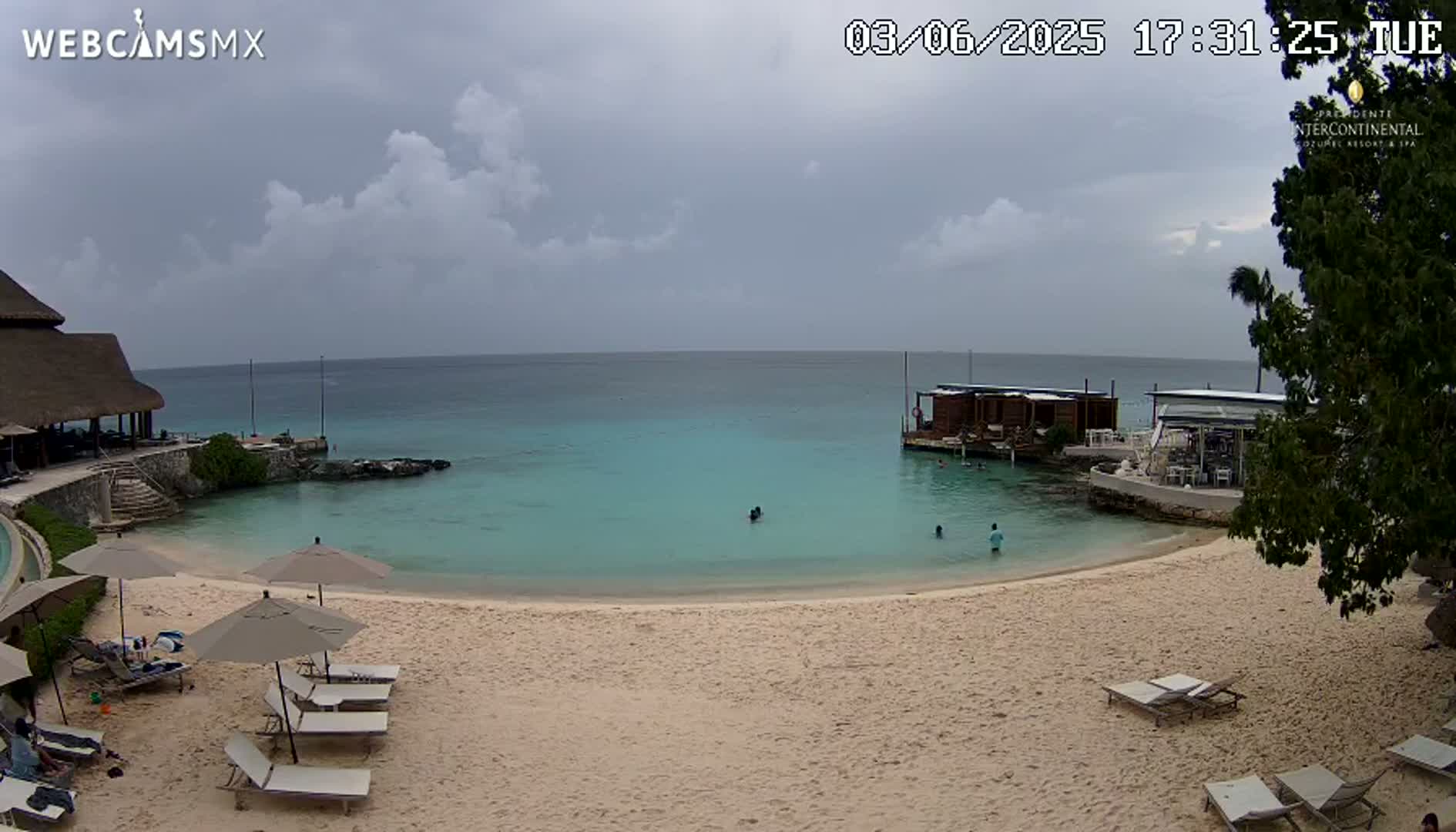 A sandy beach with lounge chairs and umbrellas overlooks calm, turquoise water, with a few people swimming near a small overwater structure, under a partly cloudy sky.
