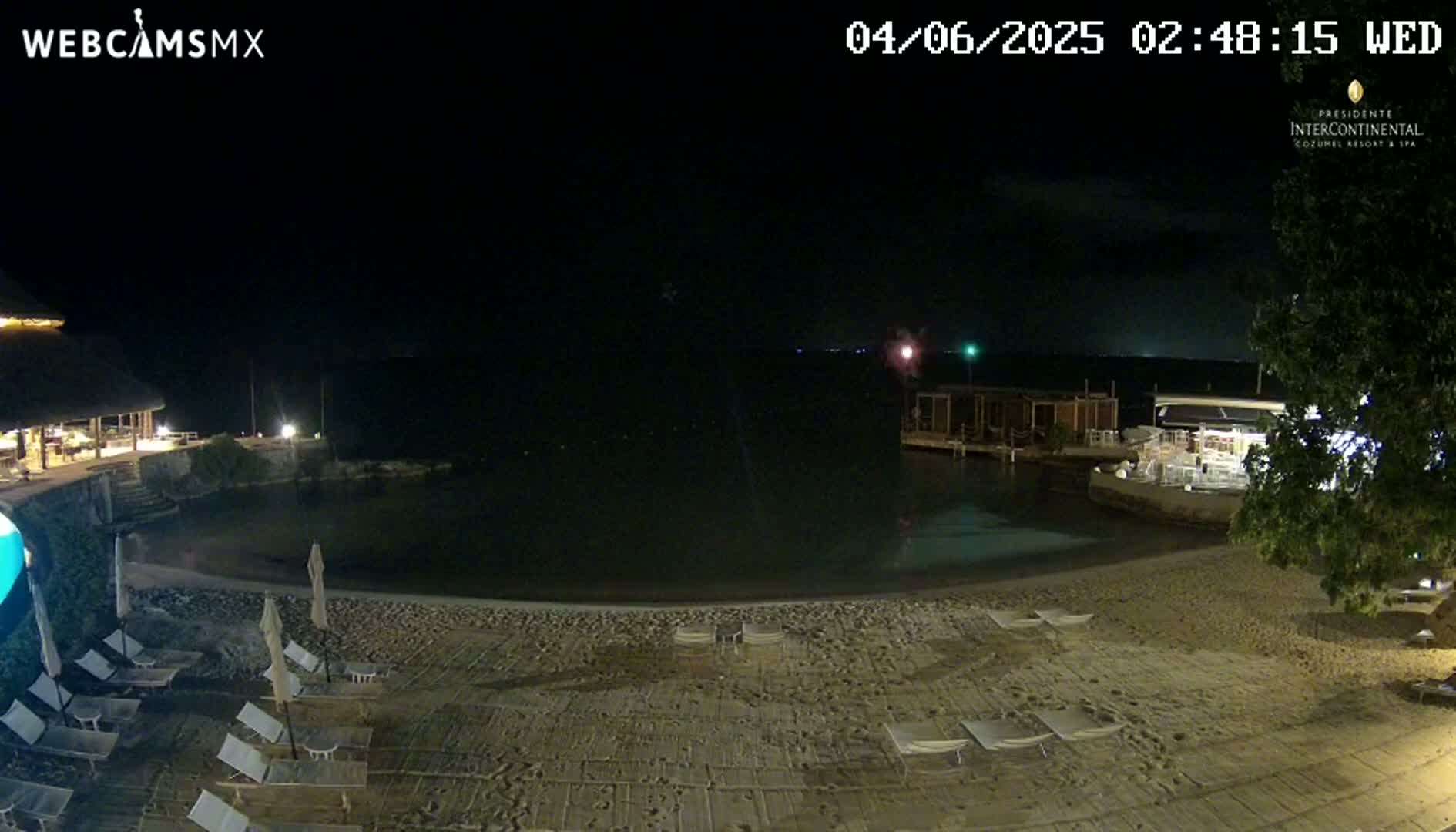 A nighttime beach scene shows a calm body of water, a sandy shoreline with empty lounge chairs, and low-lying buildings under a mostly clear, dark sky.