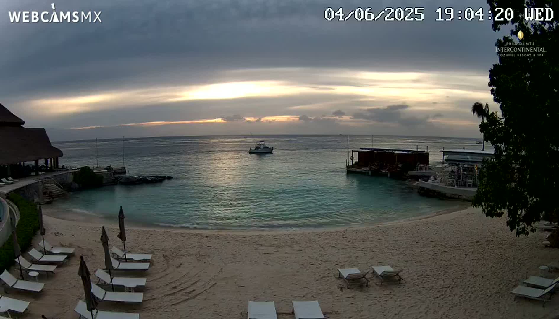 A sandy beach with lounge chairs and umbrellas sits on the shore of calm turquoise water, under an overcast sky with a pale sunset.