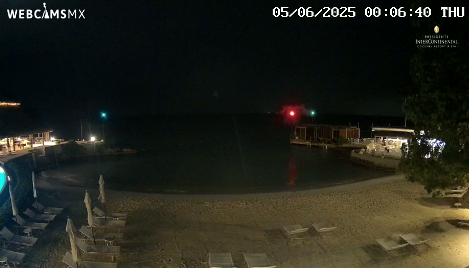 A nighttime beach scene shows a sandy shoreline with lounge chairs and umbrellas, a calm body of water, and buildings with lights in the distance under a clear, dark sky.