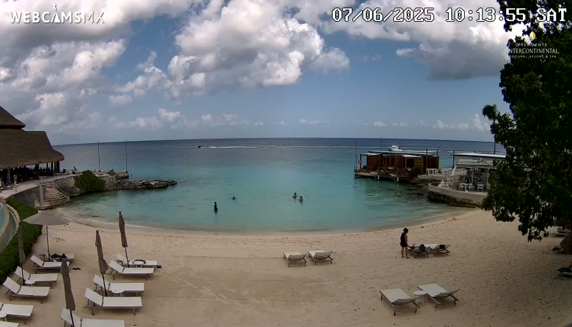 A partly cloudy day at a beach resort shows calm turquoise water, a sandy beach with lounge chairs, and a few people swimming and lounging.