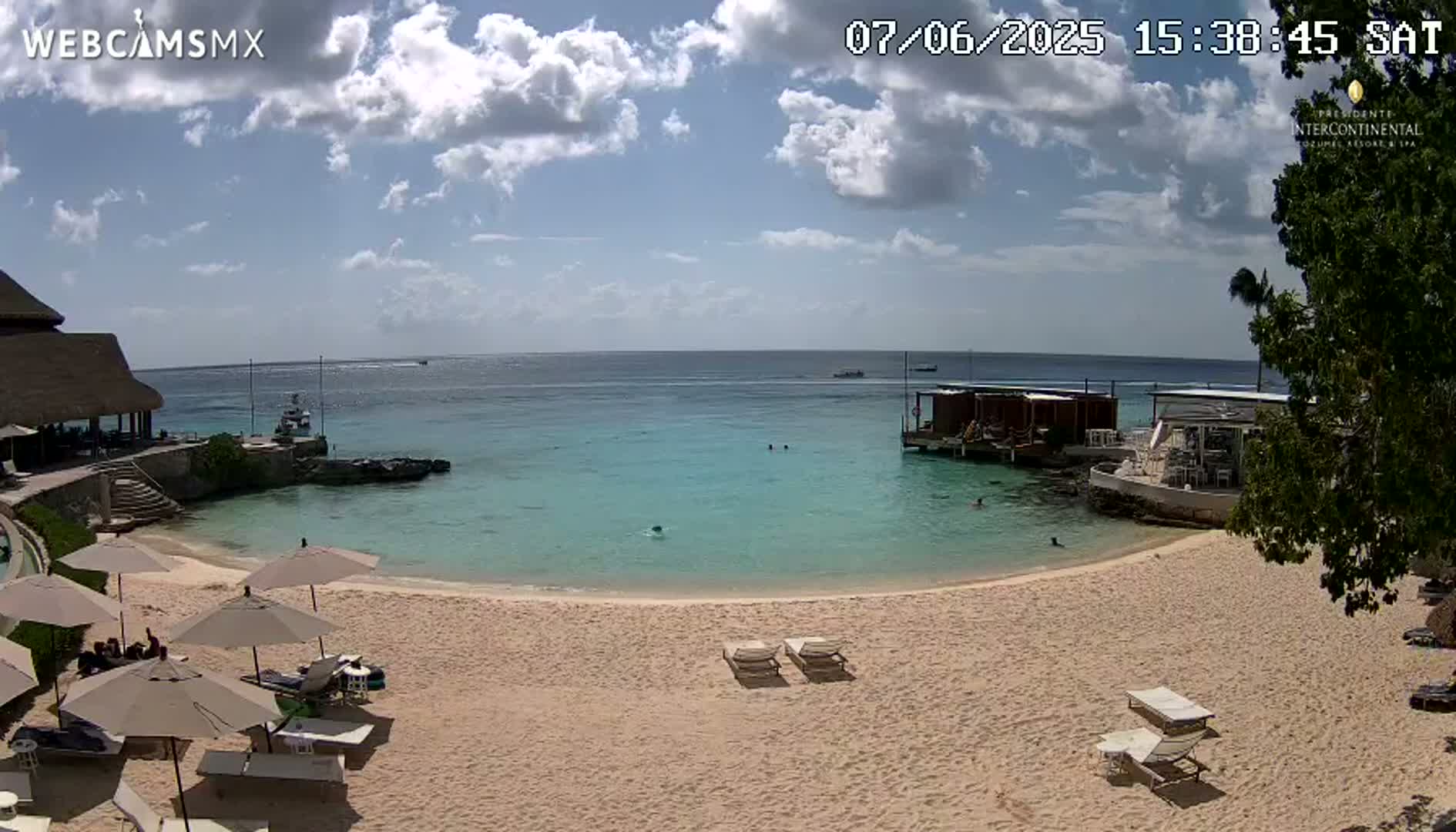 A partly sunny beach scene shows calm, turquoise water, a sandy beach with lounge chairs and umbrellas, and a building partially visible in the background.