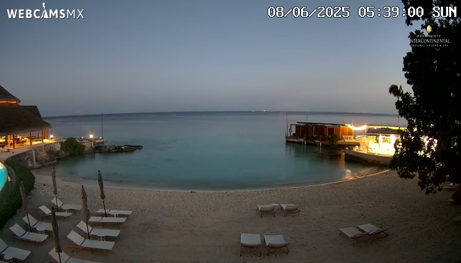 A tranquil beach scene at twilight shows a calm, turquoise ocean, a sandy beach with lounge chairs, and a partially illuminated resort building.