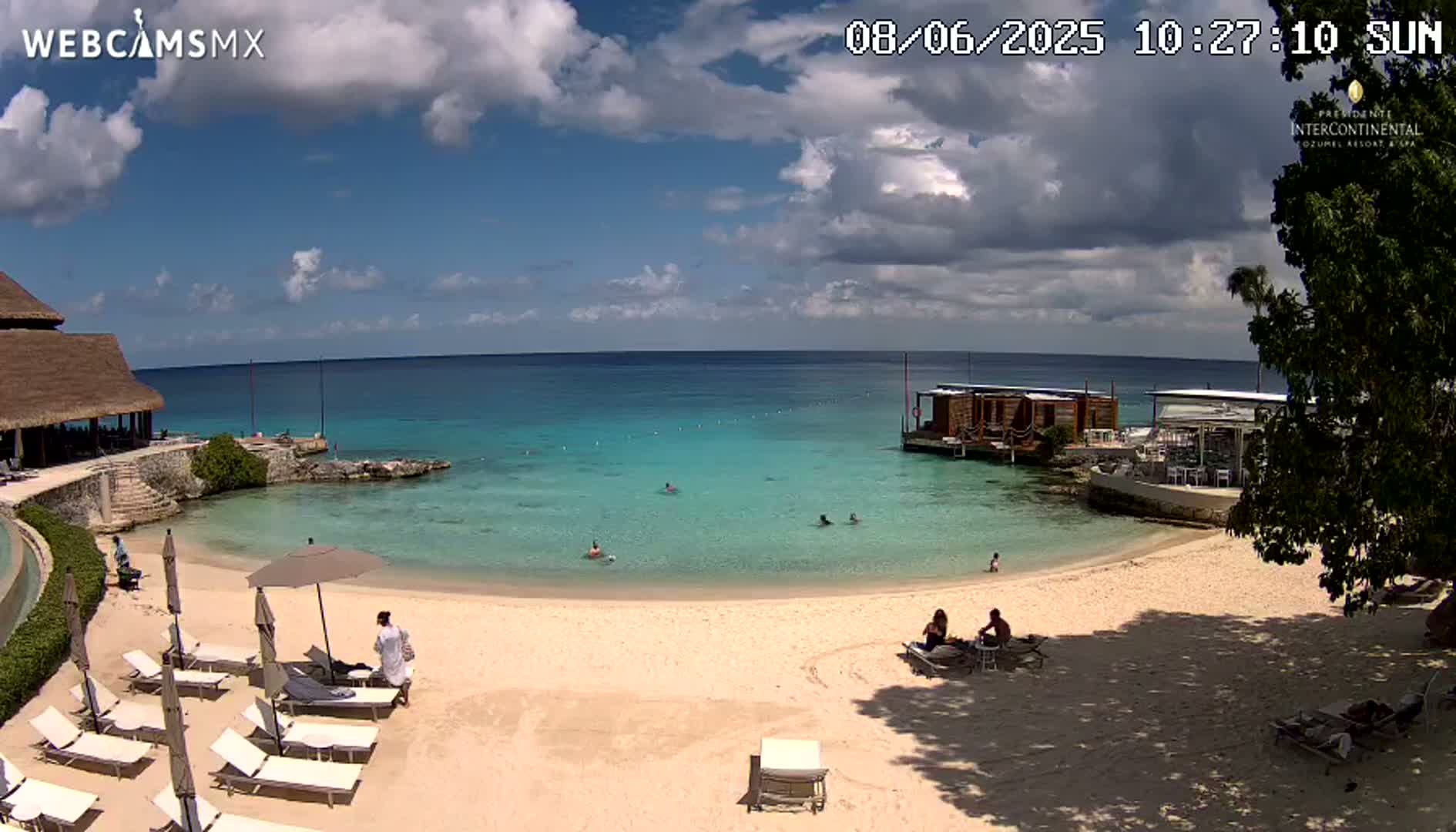 A partly cloudy sunny day at a beach with calm, turquoise water, a sandy shore, lounge chairs, people swimming and relaxing, and buildings along the shoreline.