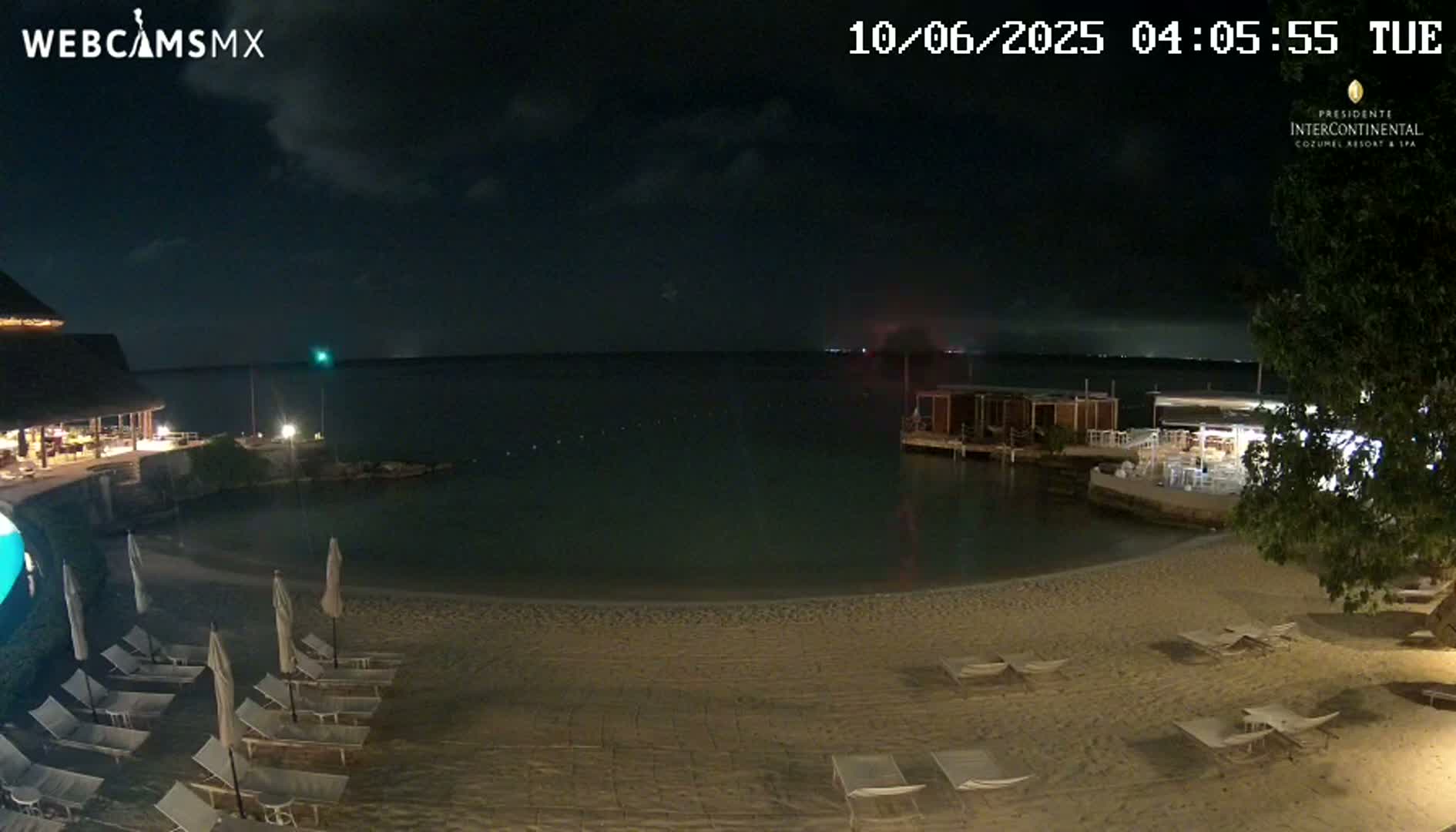 A nighttime view of a calm, dark body of water with a sandy beach lined with lounge chairs, a few buildings, and some sparse vegetation.