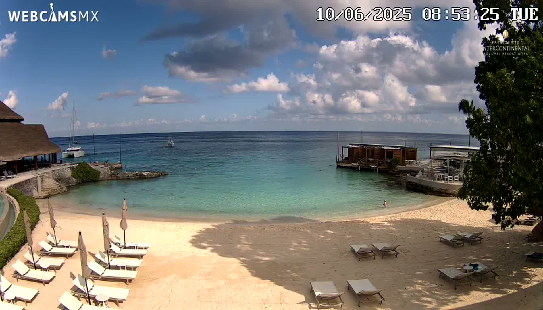 A partly sunny day at a beach resort shows calm, turquoise water, a sandy beach with lounge chairs and umbrellas, and a building partially in the water.