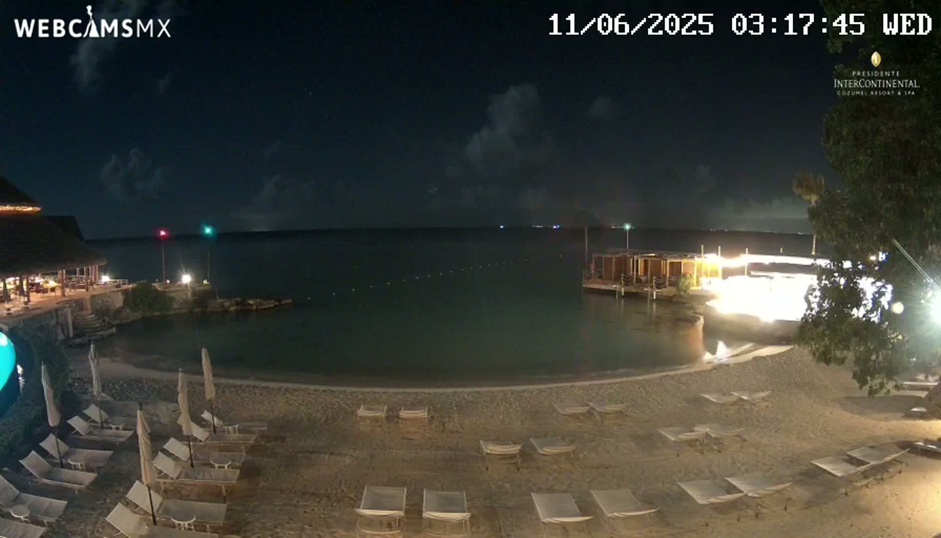 A nighttime beach scene with calm water, a sandy shore lined with lounge chairs, and a building with lights visible in the background under a partly cloudy sky.