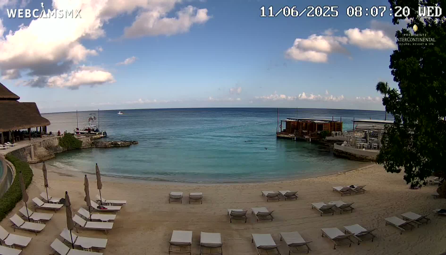 A beach with lounge chairs and umbrellas, a calm ocean with a small structure in the water, and a partially cloudy sky.