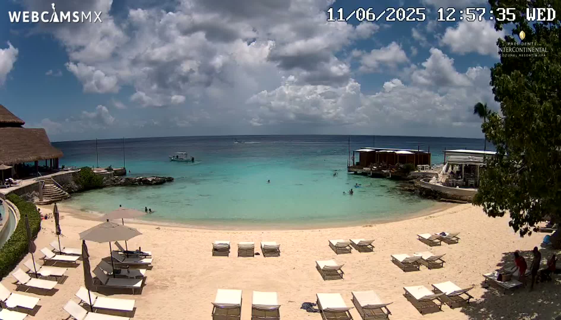 A partly cloudy day at a beach with turquoise water, a sandy shore lined with lounge chairs and umbrellas, and several small buildings on the water's edge.