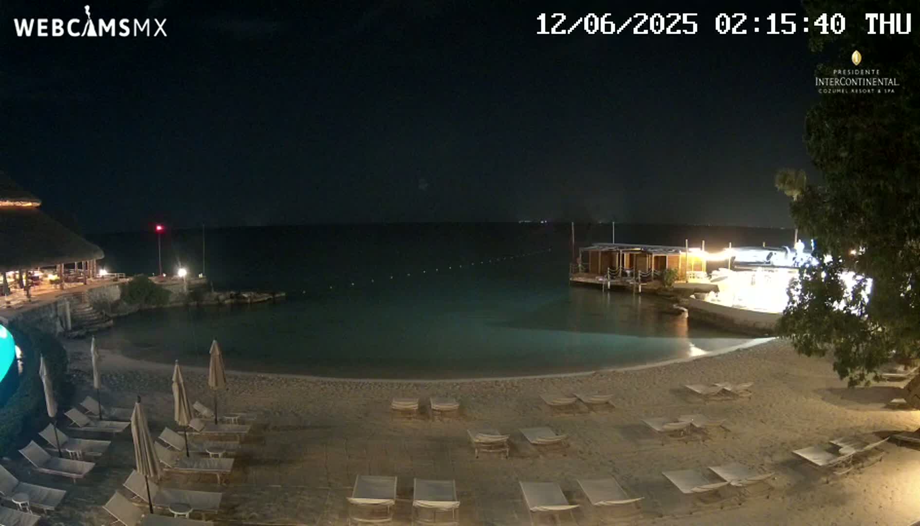 A nighttime beach scene shows a calm body of water, a sandy shore with lounge chairs and umbrellas, and a building with a thatched roof in the background under a clear, dark sky.