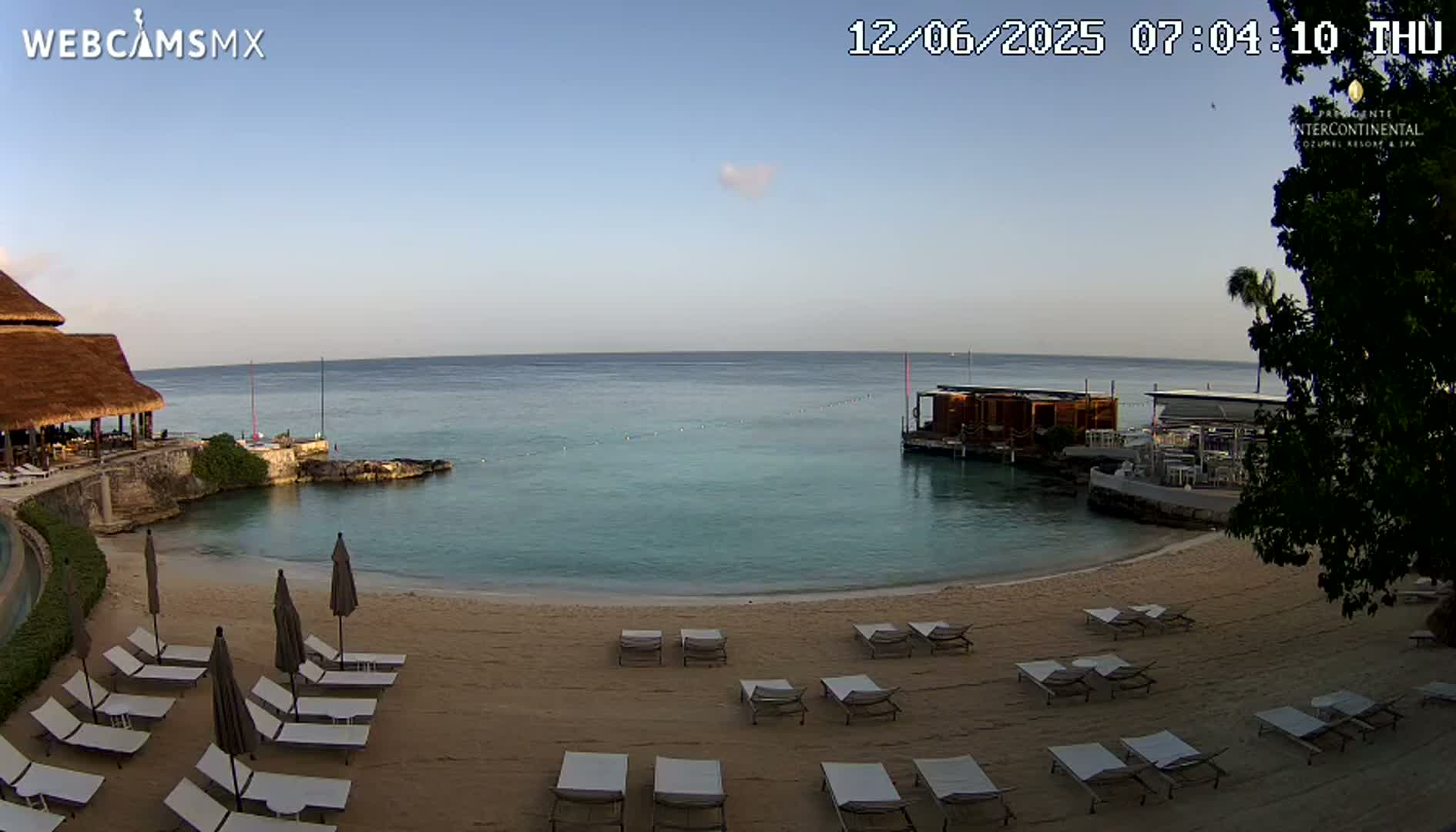 A calm, clear day shows a sandy beach with lounge chairs and umbrellas, overlooking a calm turquoise ocean with a small overwater structure and a building with a thatched roof.