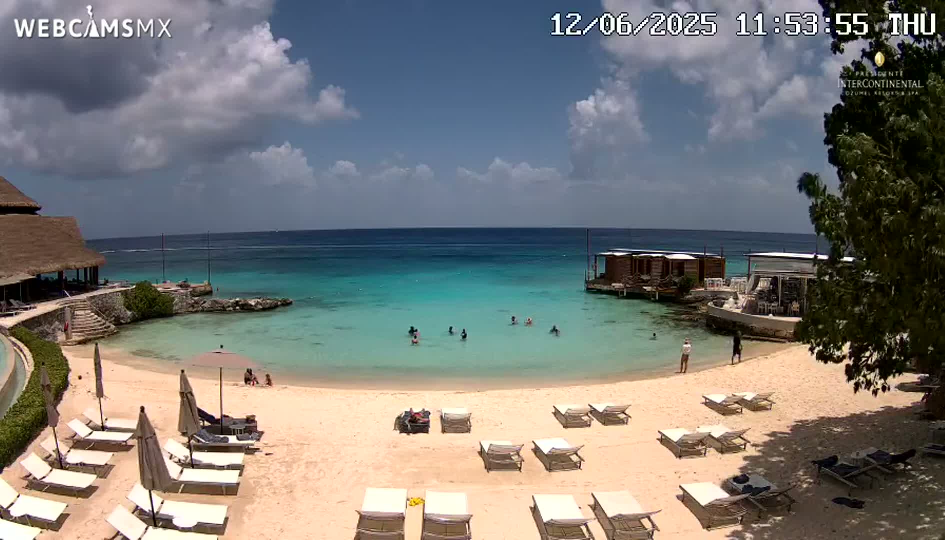 A partly cloudy sunny day at a beach resort with turquoise water, several people swimming, and lounge chairs set up on a sandy beach.