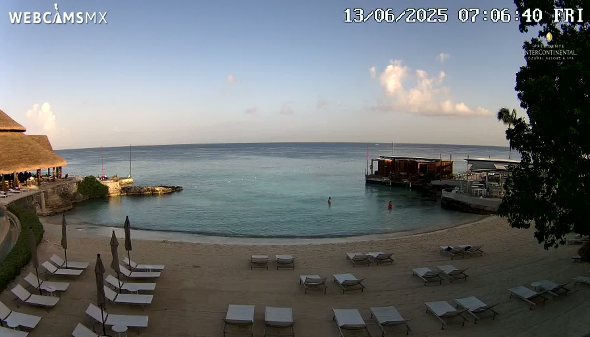 A sandy beach with lounge chairs and umbrellas sits adjacent to calm, clear water with a few people swimming, under a mostly clear sky with a few clouds.