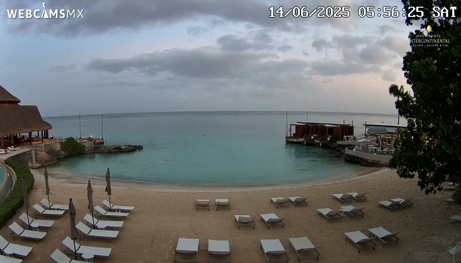 A mostly cloudy dawn scene shows a sandy beach with lounge chairs and umbrellas, overlooking calm turquoise water and a resort building on a small peninsula.