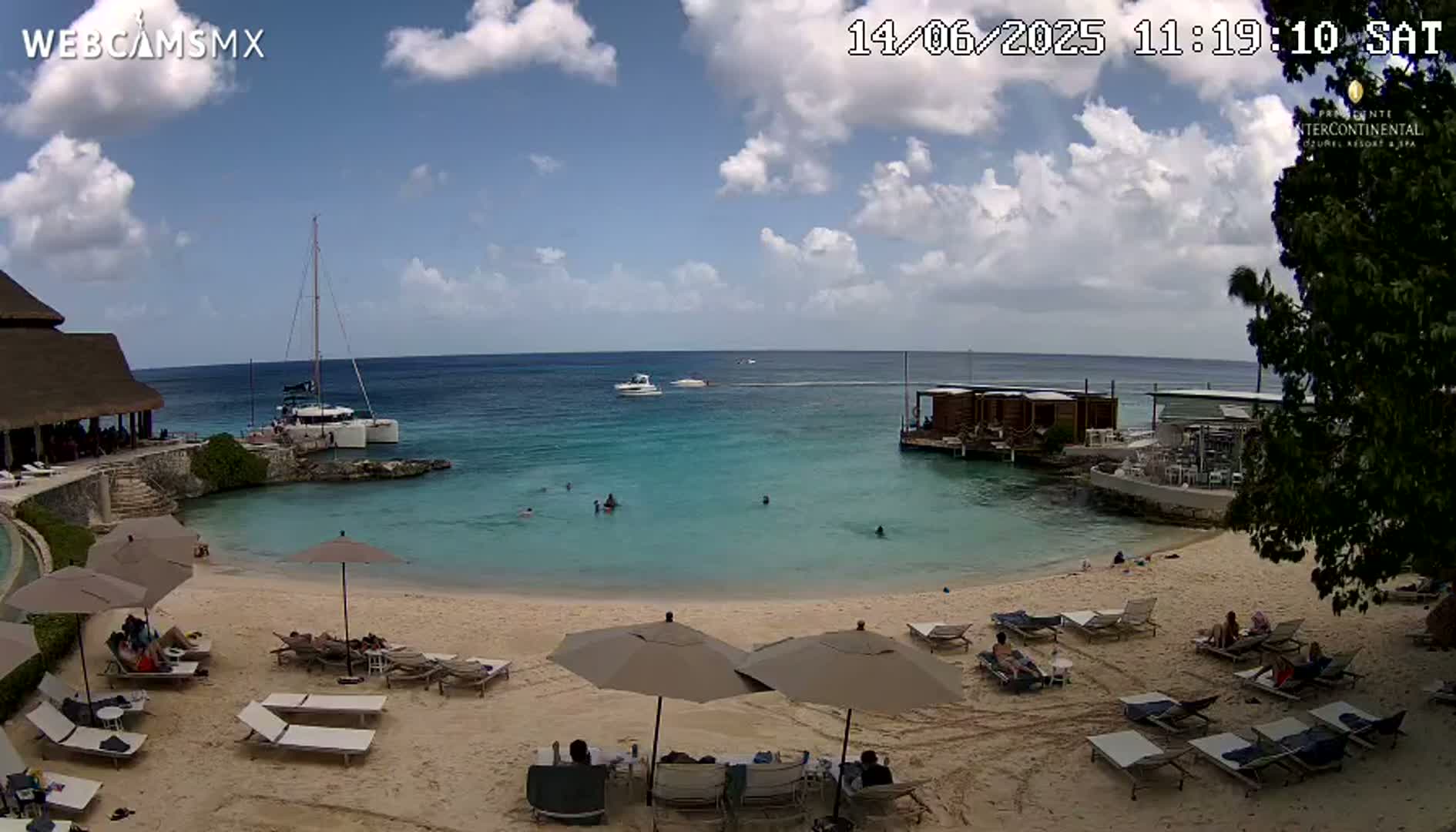 A partly cloudy day at a beachside resort shows a sandy beach with lounge chairs and umbrellas, calm turquoise water with several people swimming, and boats moored offshore.
