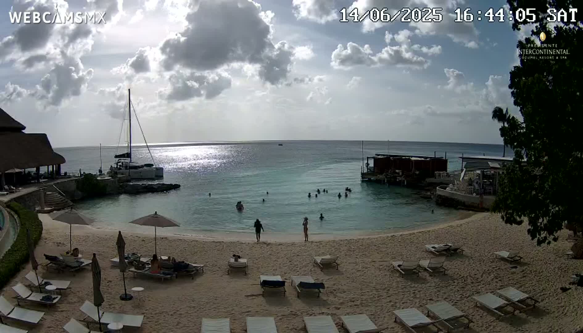 A partly cloudy day at a beachside resort shows a sandy beach with lounge chairs and umbrellas, calm turquoise water with several people swimming, and boats moored offshore.